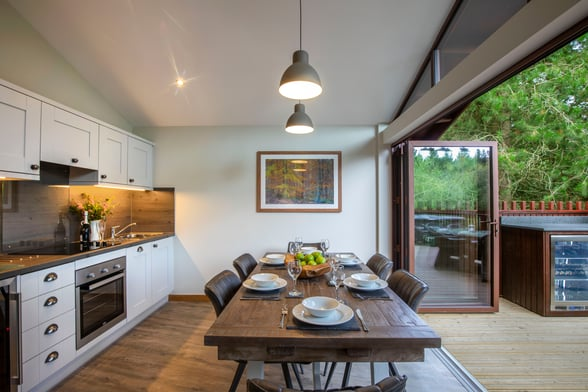 Modern dining area with a wooden table set for six, large windows showcasing greenery, and a stylish kitchen in Yorkshire.