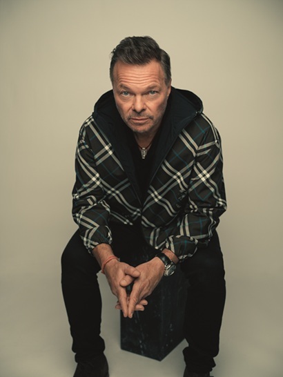 Danny Rampling seated on a stool, wearing a checkered jacket, with a serious expression against a neutral backdrop.
