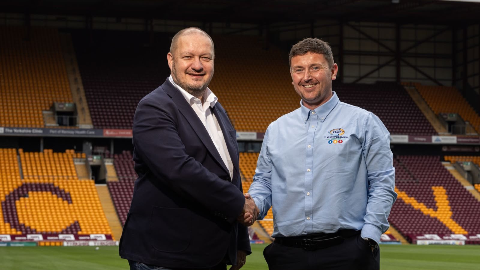 Two men shaking hands on a football pitch with stands behind