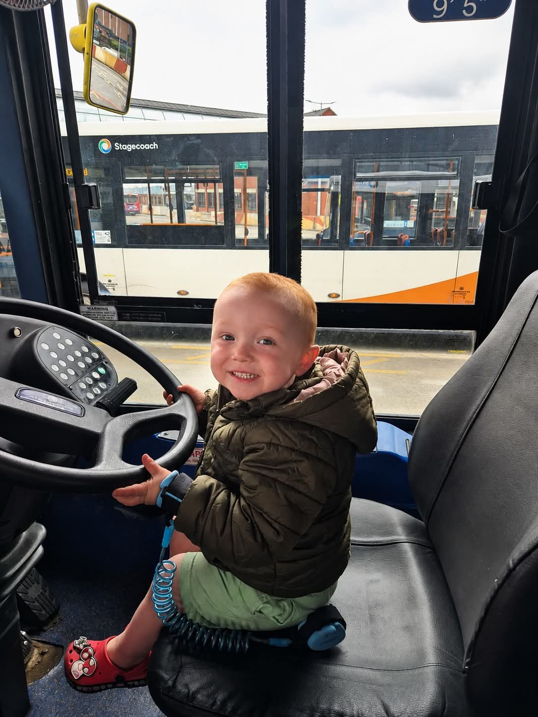 Smiling child in a bus driver's seat with another bus outside