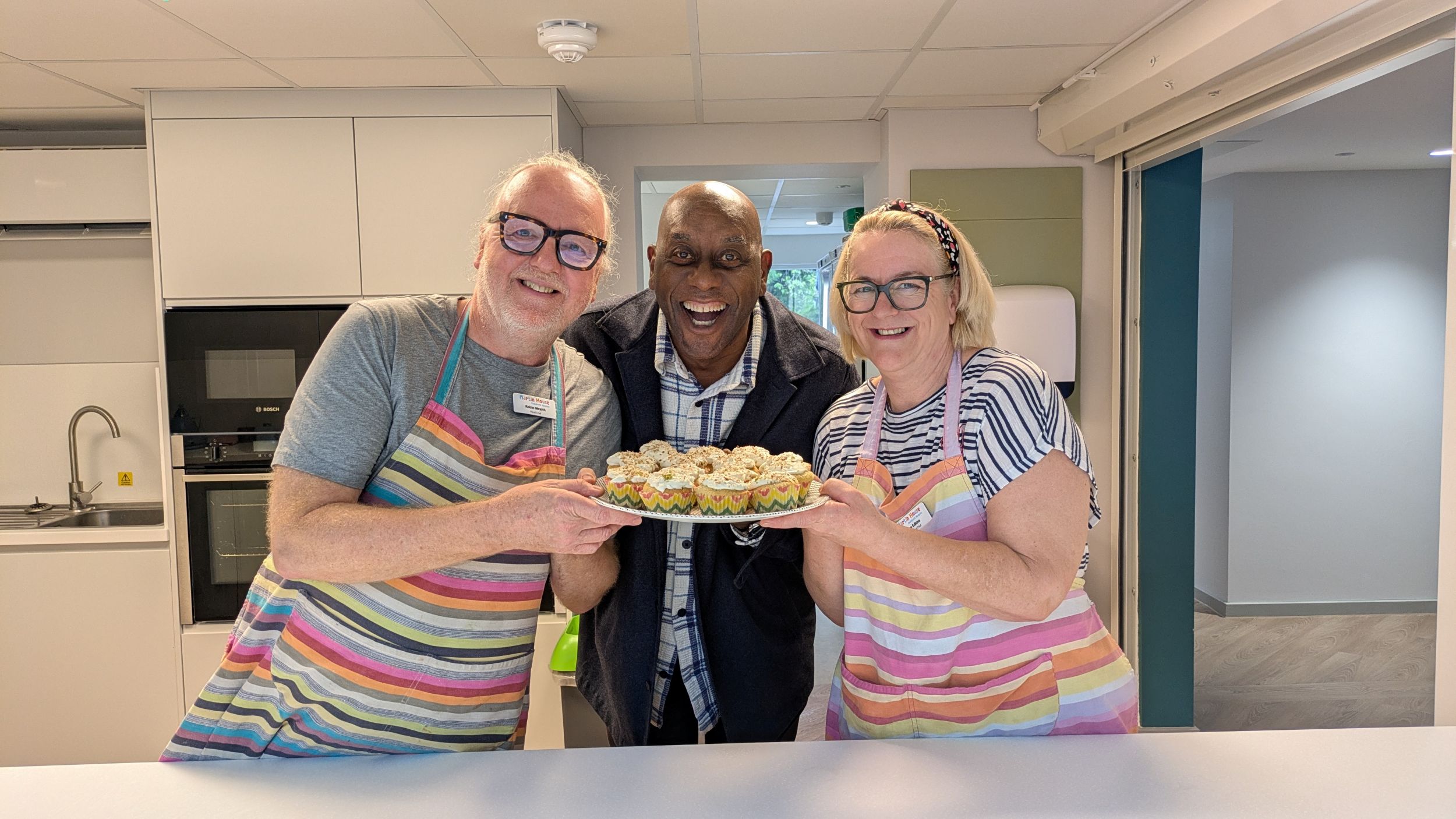 Three smiling people in aprons presenting a tray of cupcakes in a modern kitchen