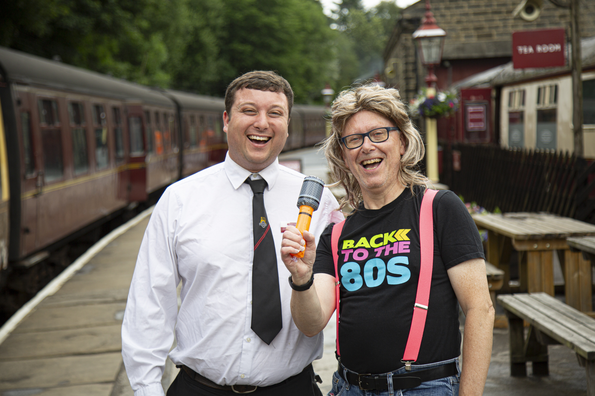 Two men smiling on a vintage train platform, one holds a microphone