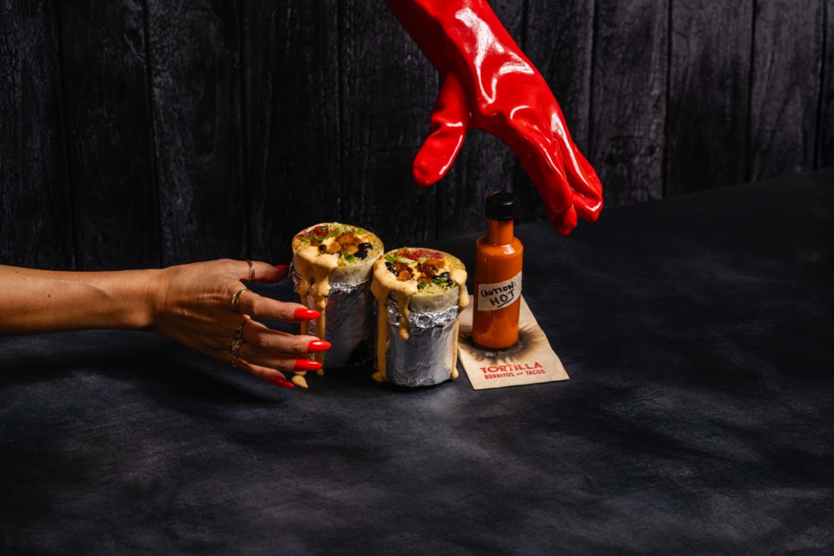 Two burritos with hot sauce bottle on dark table, red gloves