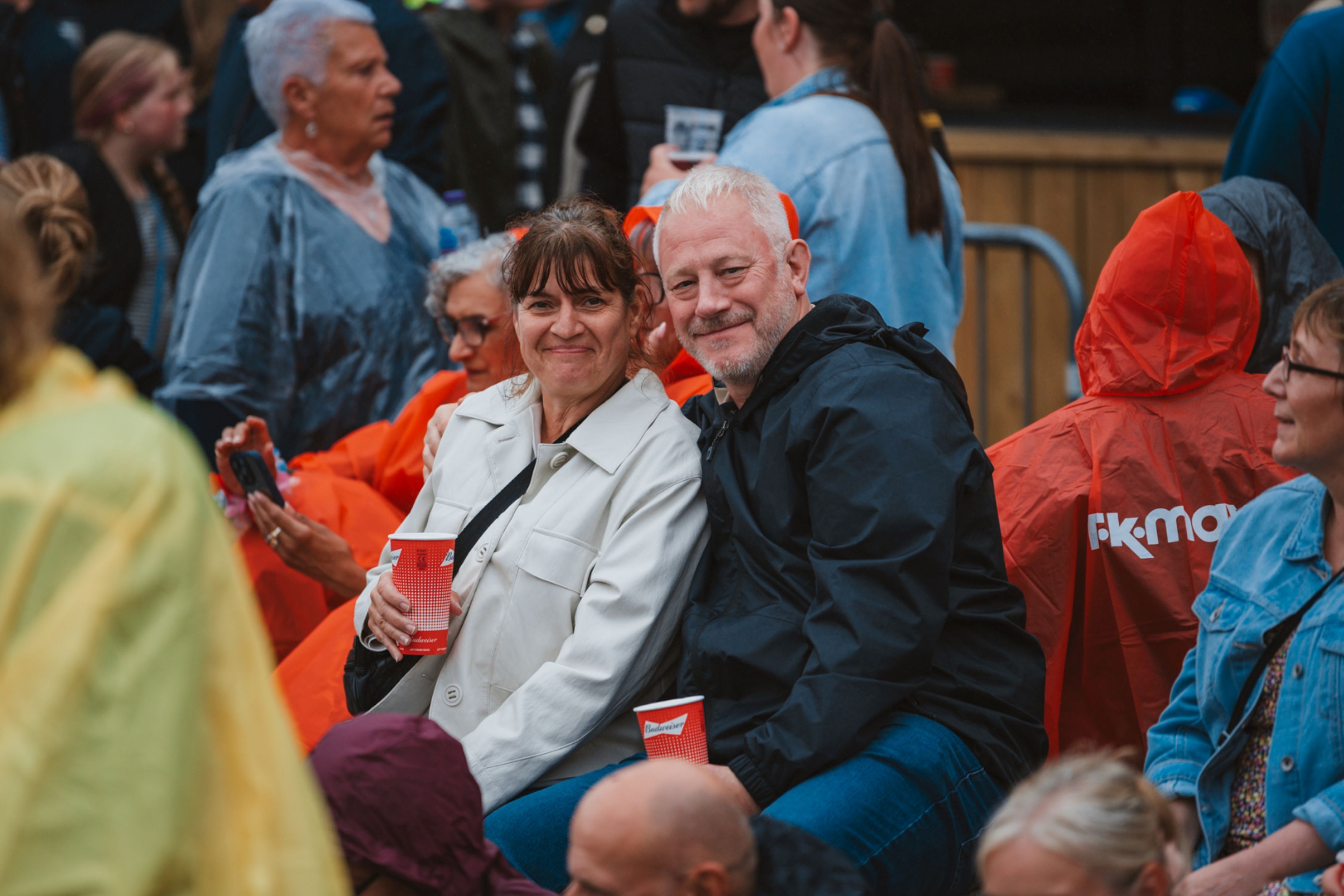 Smiling couple in raincoats at outdoor event amid crowd