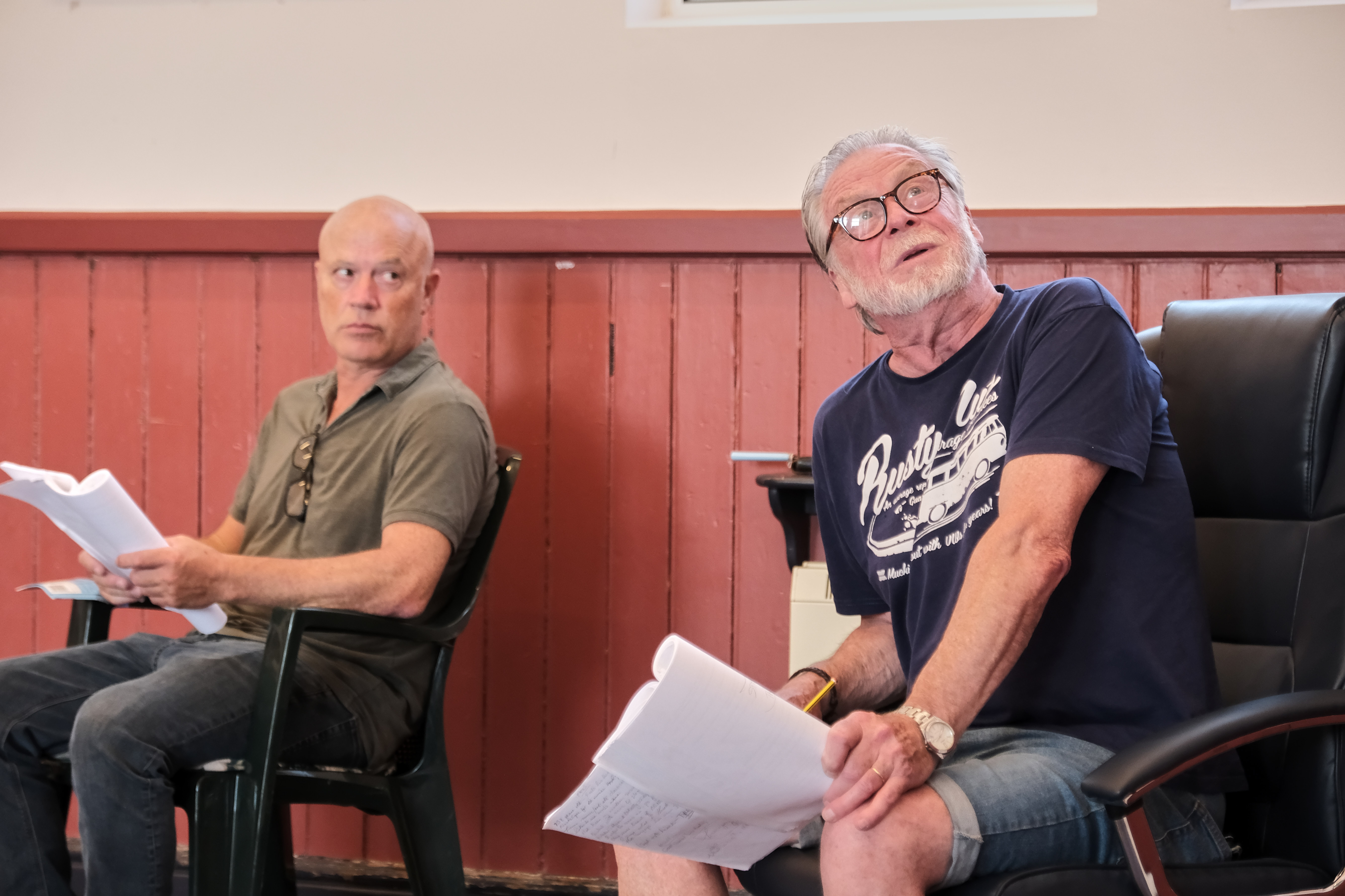 Two men seated indoors reading scripts against red wall