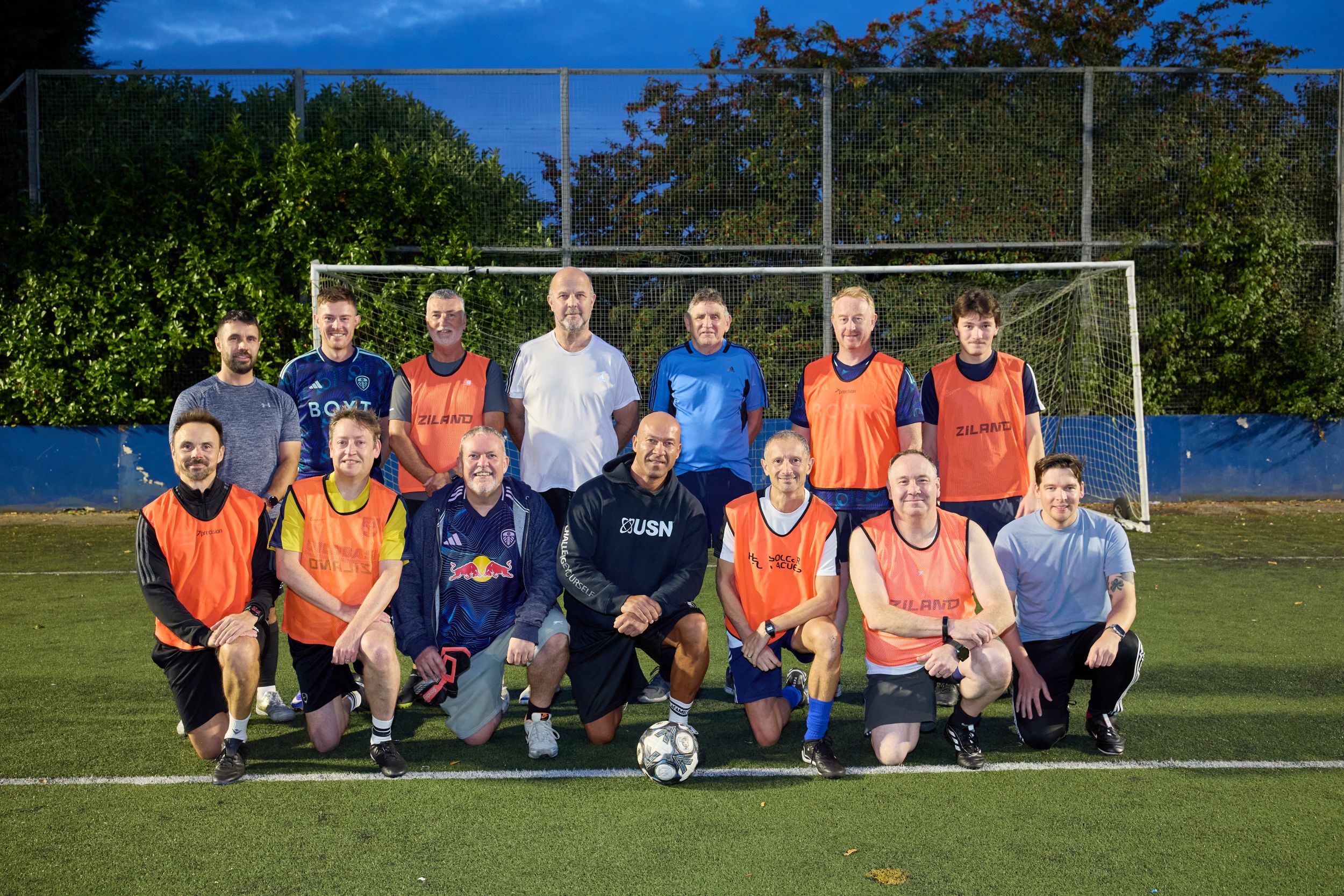 Group of men in sports kits posing on a football pitch at dusk