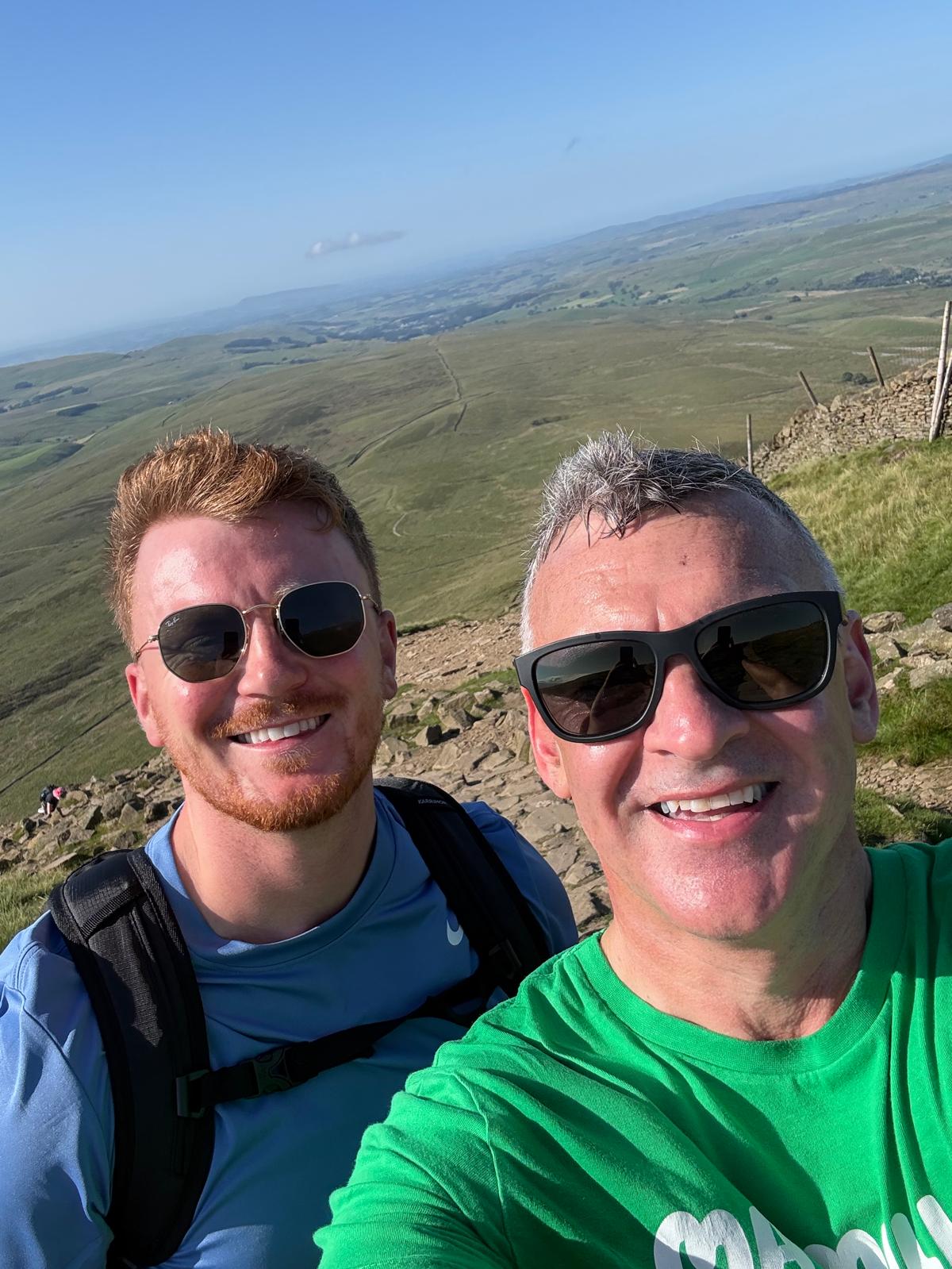 Two smiling hikers with sunglasses on a sunny peak overlooking green hills