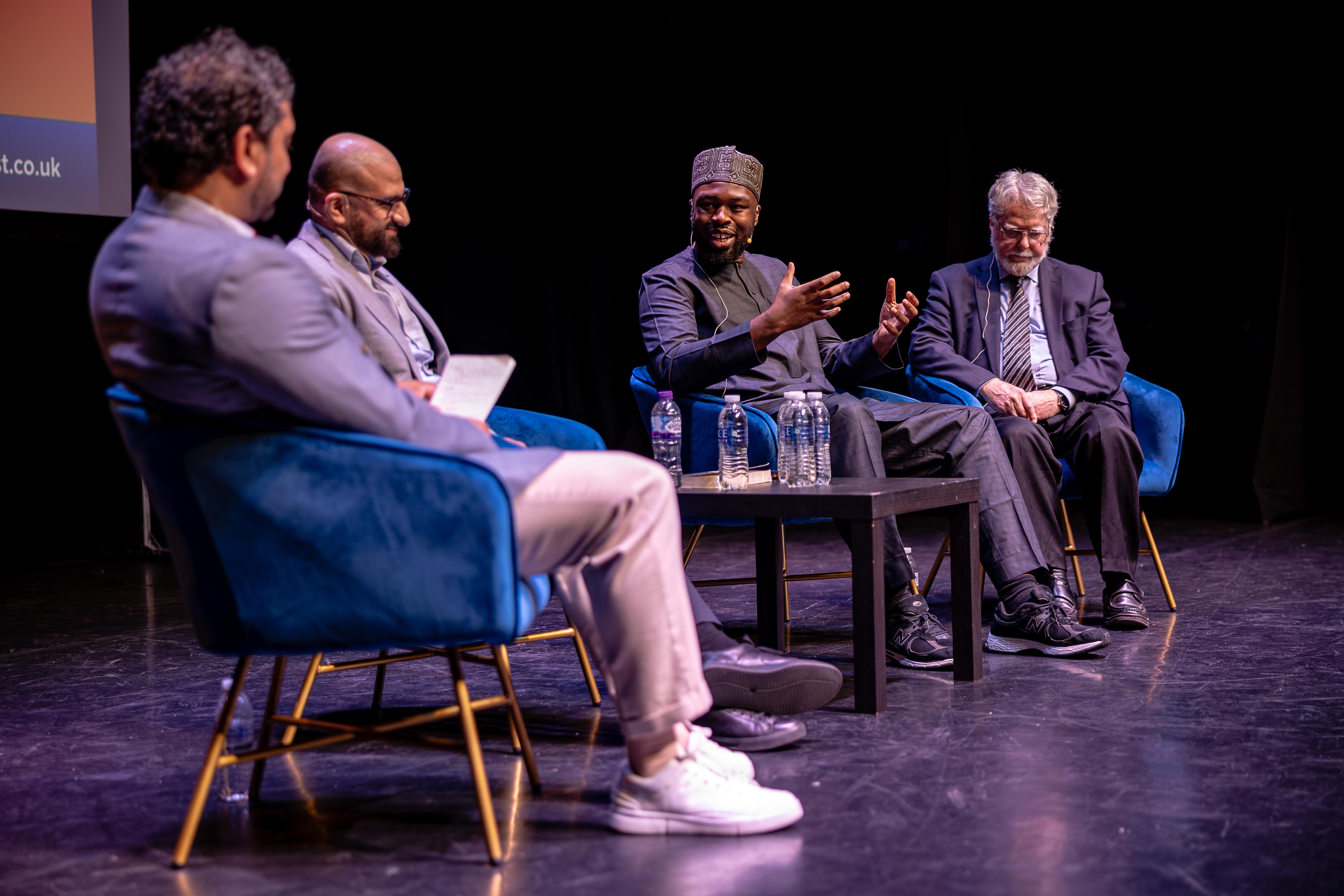 Four men in discussion on stage with blue chairs and dark backdrop
