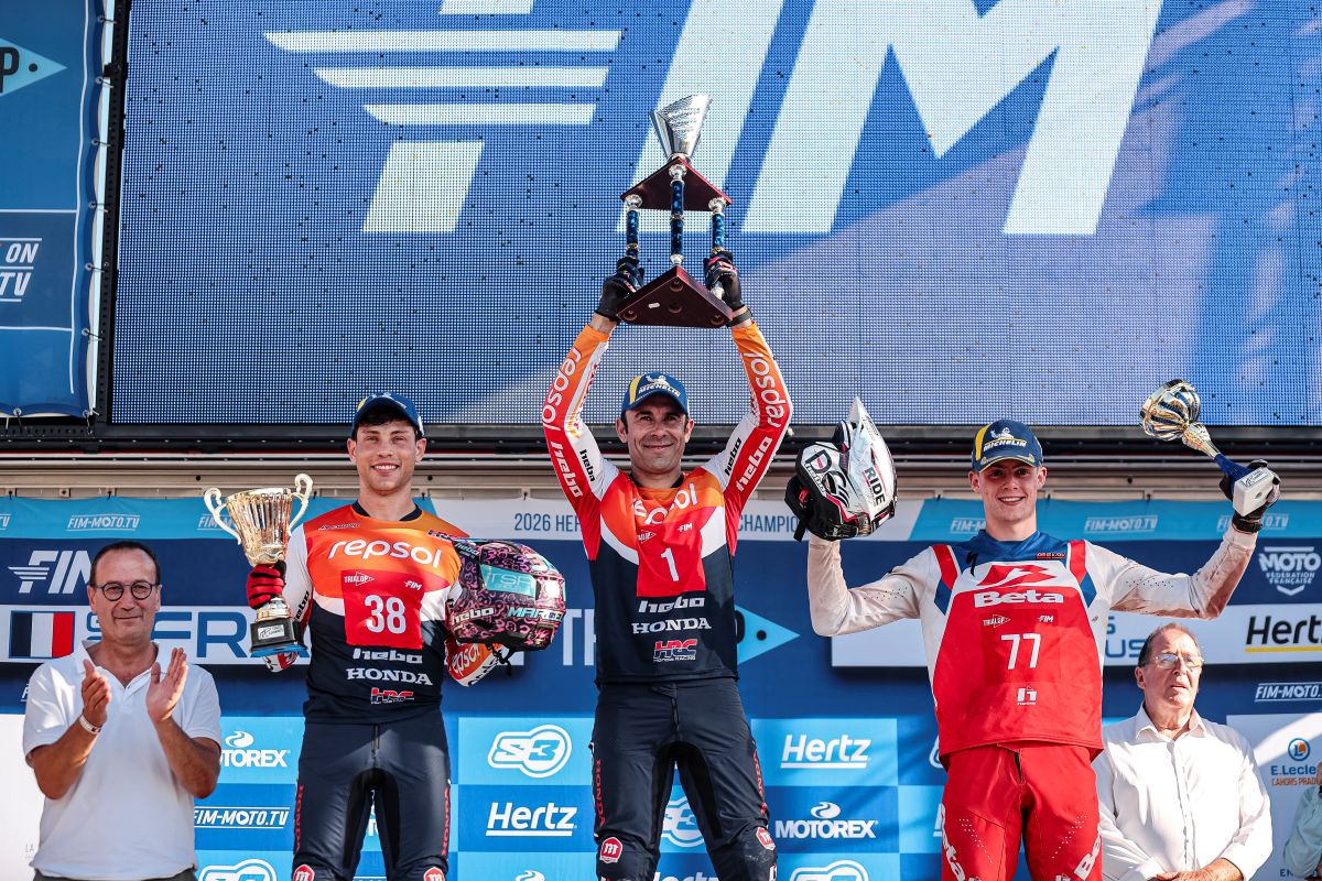 Three motorbike racers holding trophies on a podium with a backdrop