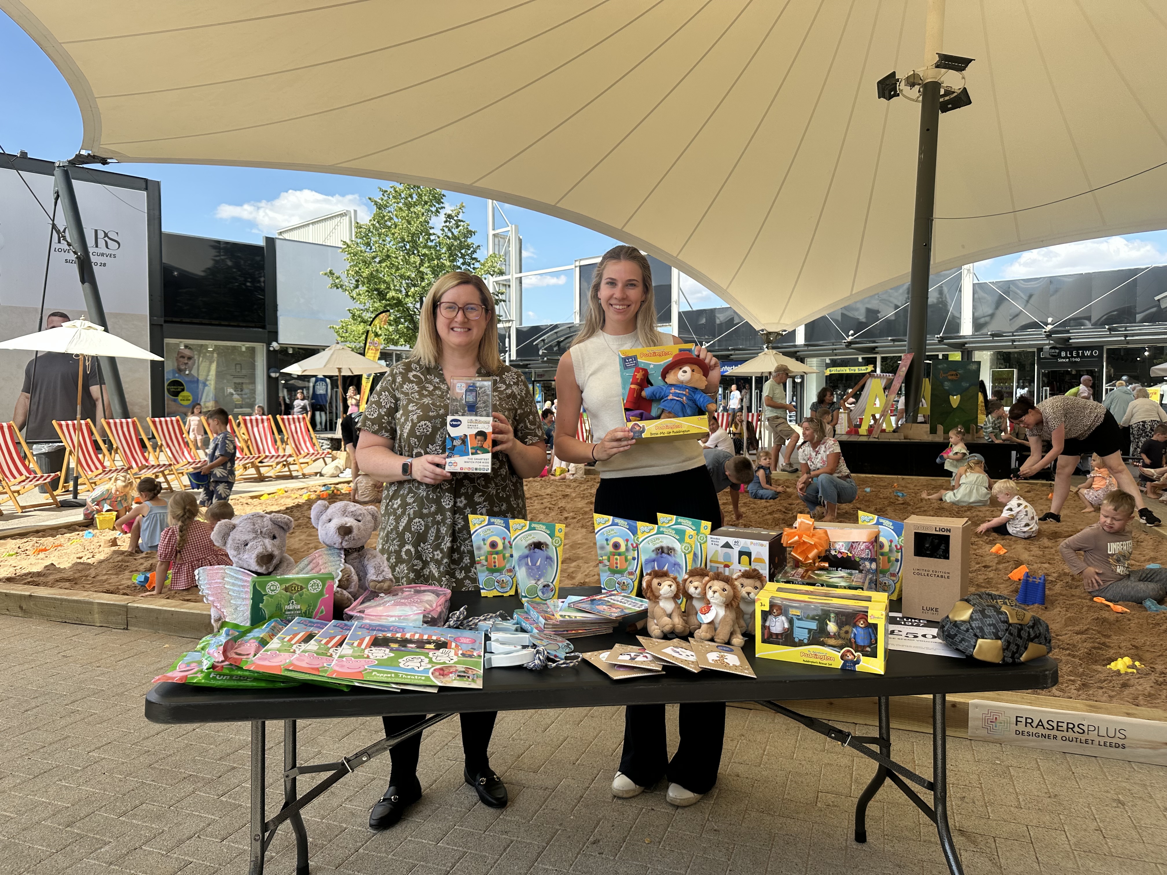 Two women showcasing toys at an outdoor market under a canopy