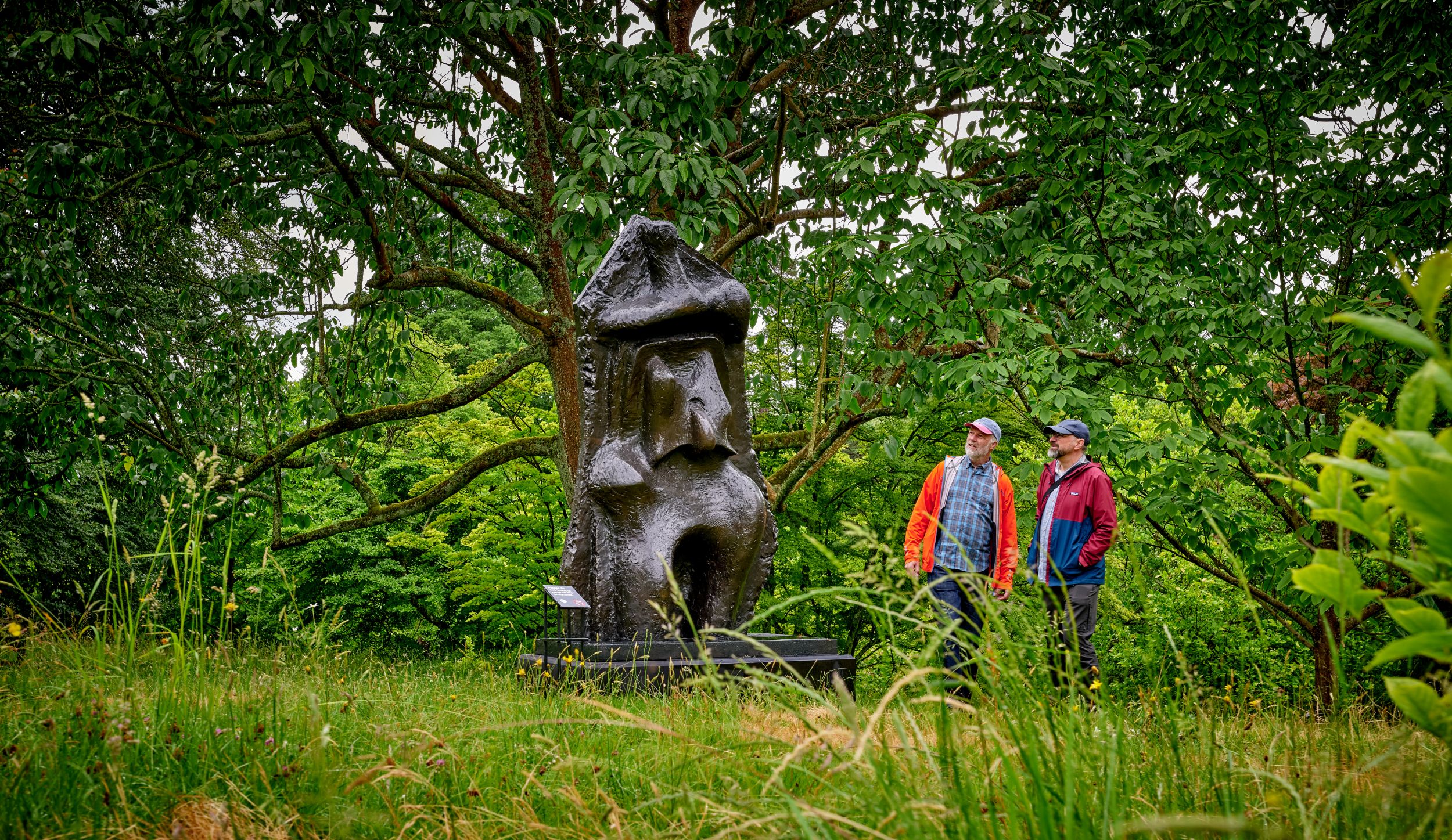 Two people admiring a large abstract stone sculpture in a lush park