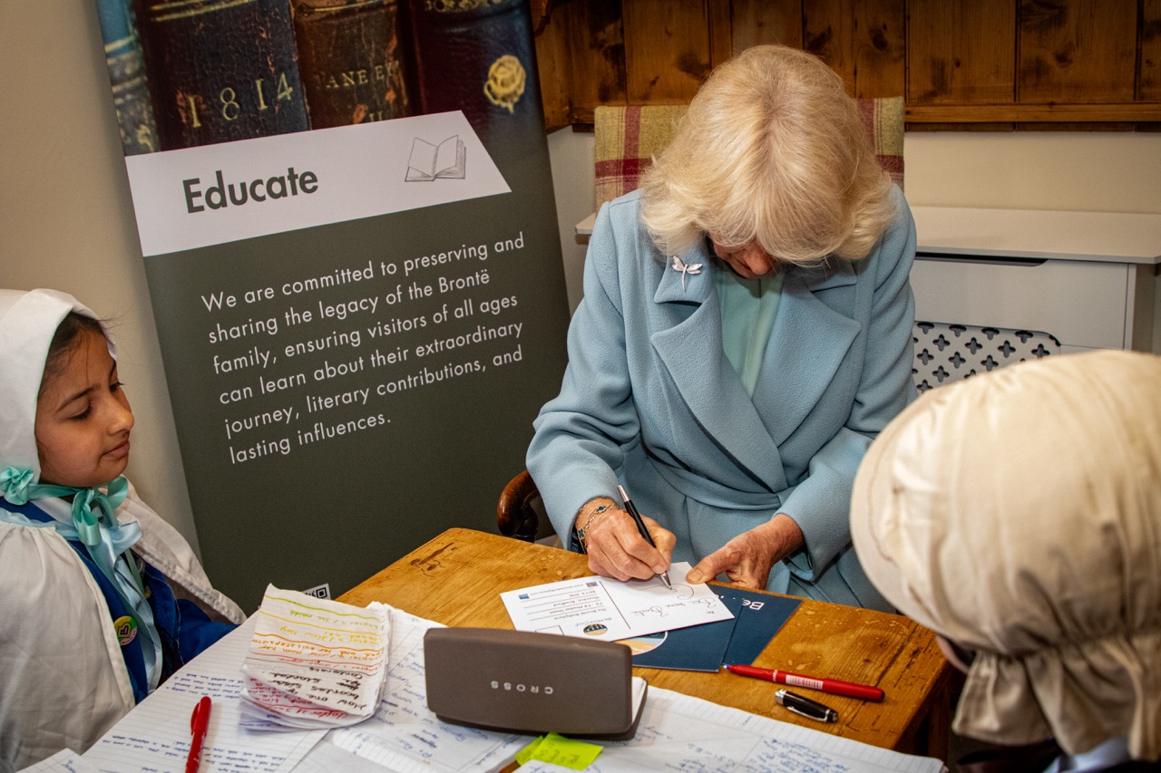 HM Queen Camilla writing at table during educational event