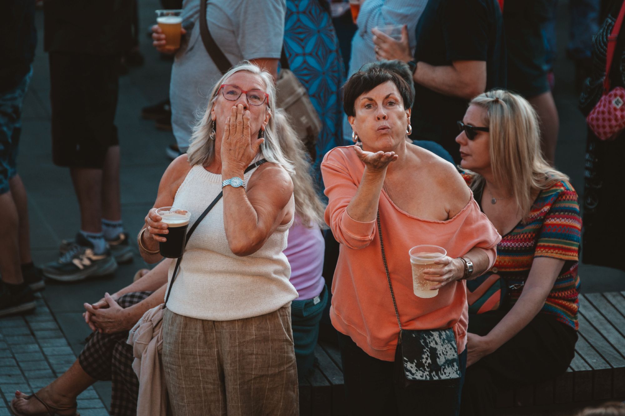 Two women enjoying a street festival, blowing kisses with drinks in hand