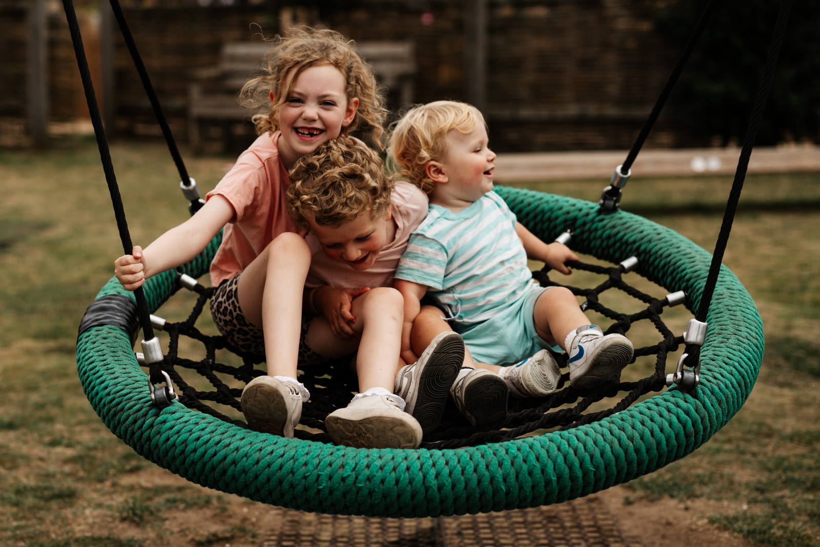 Three children laughing on a green rope swing in a park