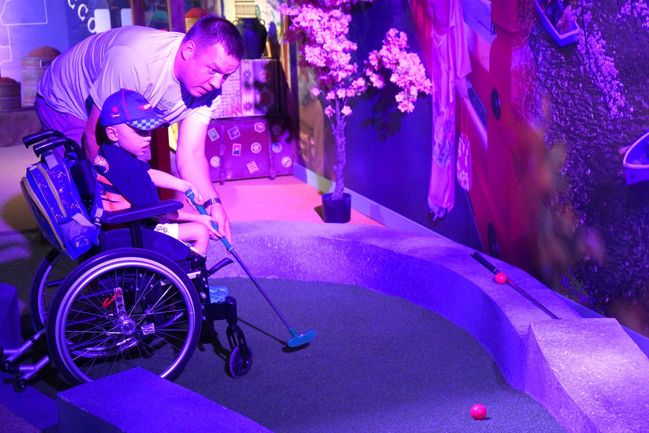 Man assisting child in wheelchair playing mini-golf indoors