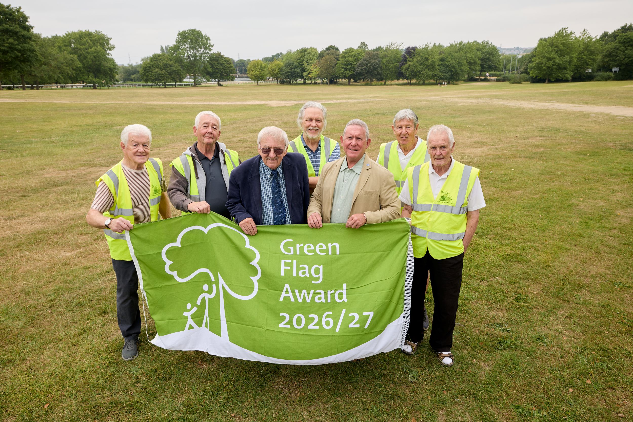 Group of seven elderly men in high-vis vests holding Green Flag Award banner on grassy field