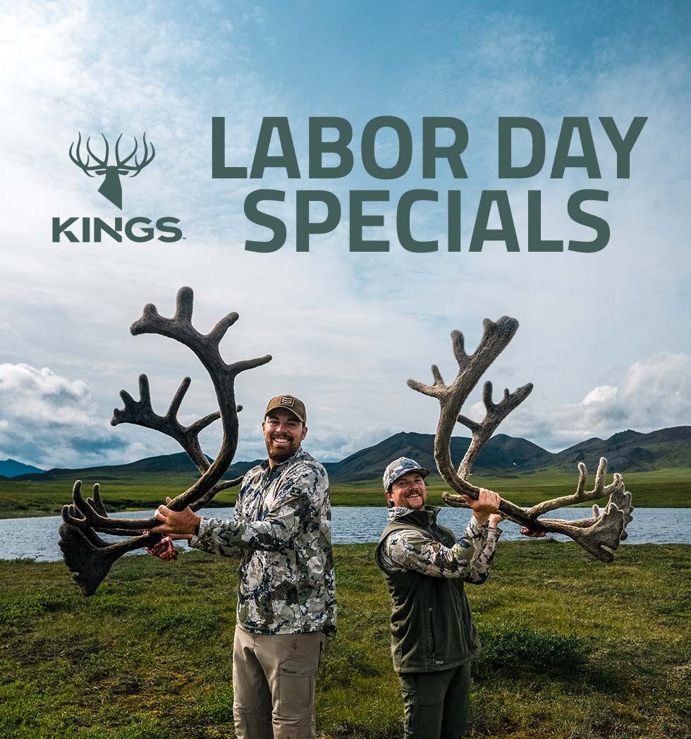 Two people in camouflage holding large antlers in open landscape