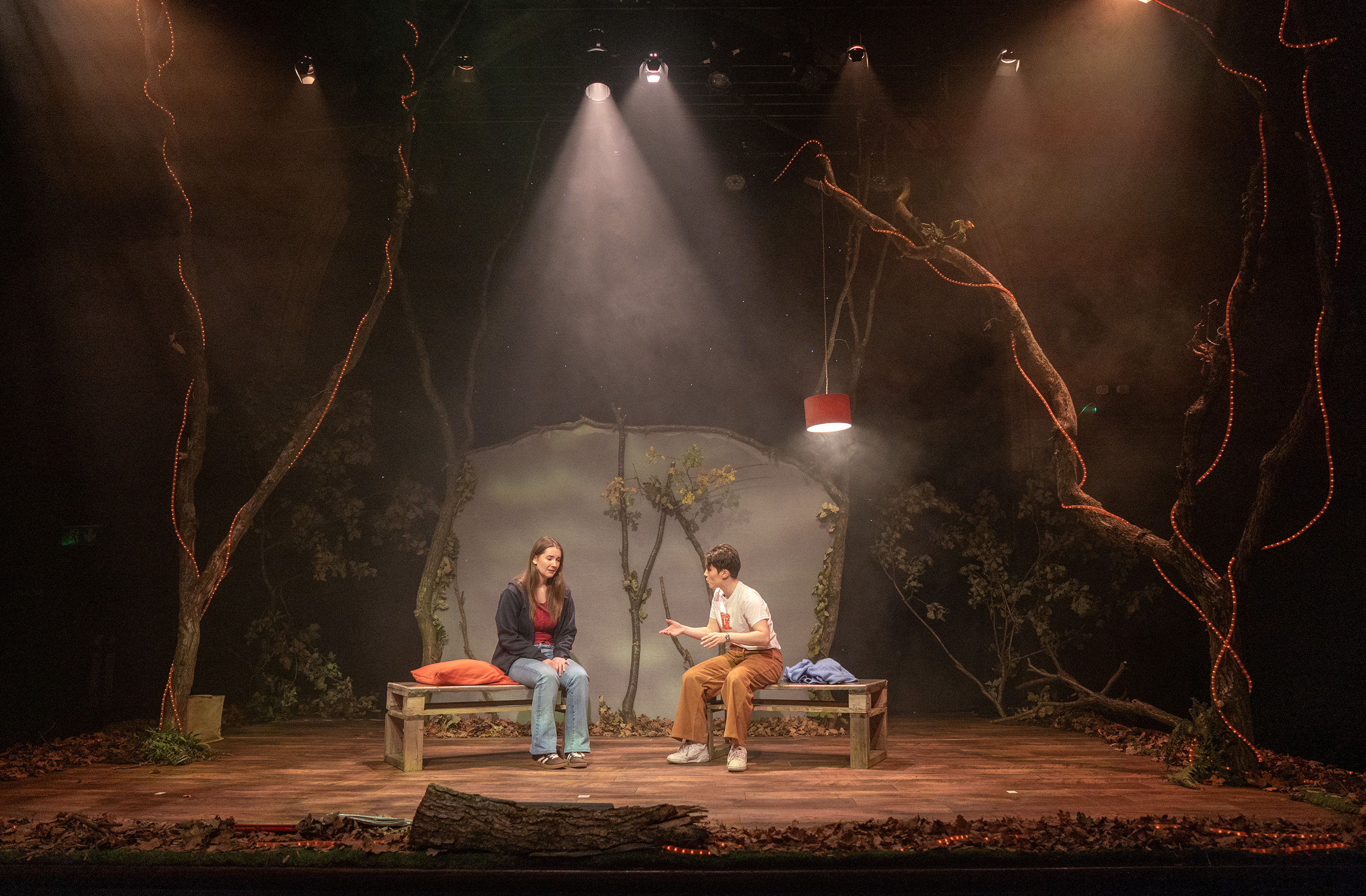 Two actors on stage surrounded by woodland set under spotlight