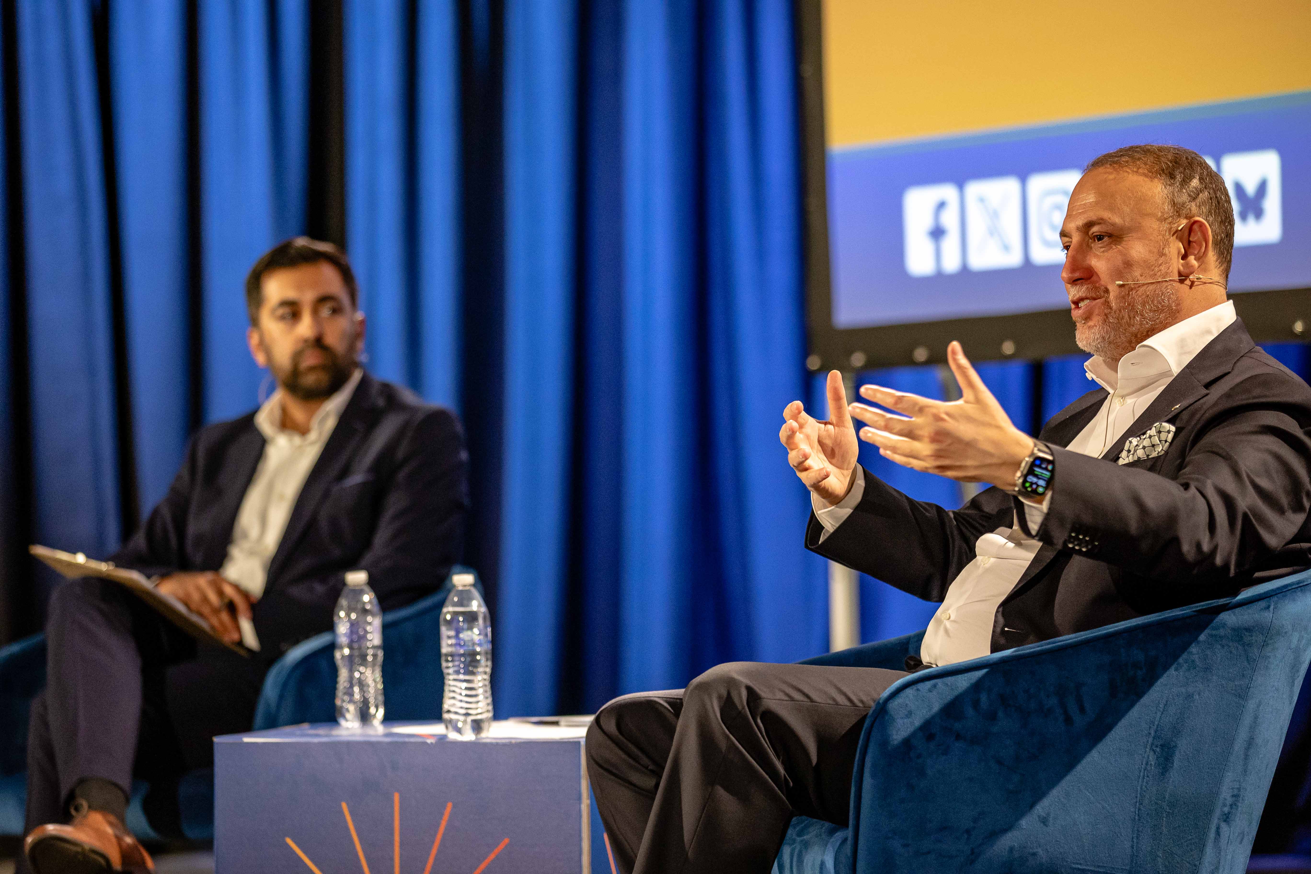 Two men in suits engaged in discussion on stage with blue curtains