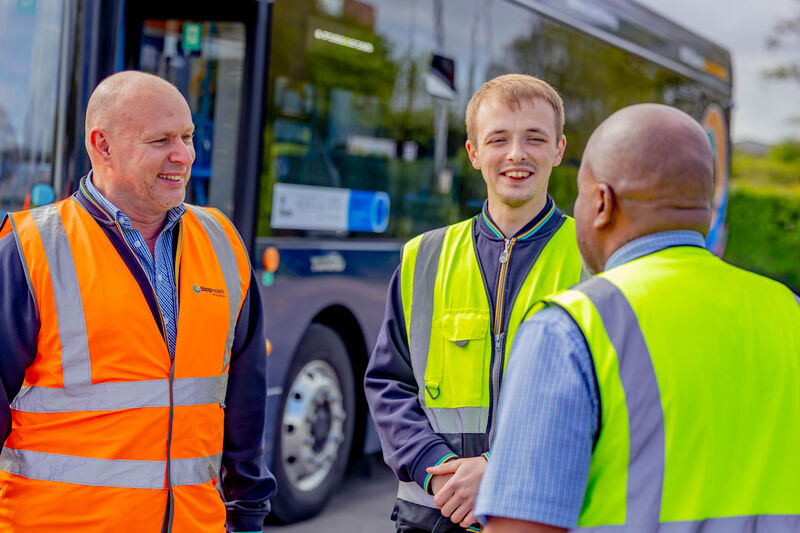 Three bus drivers in high-visibility vests chatting outdoors