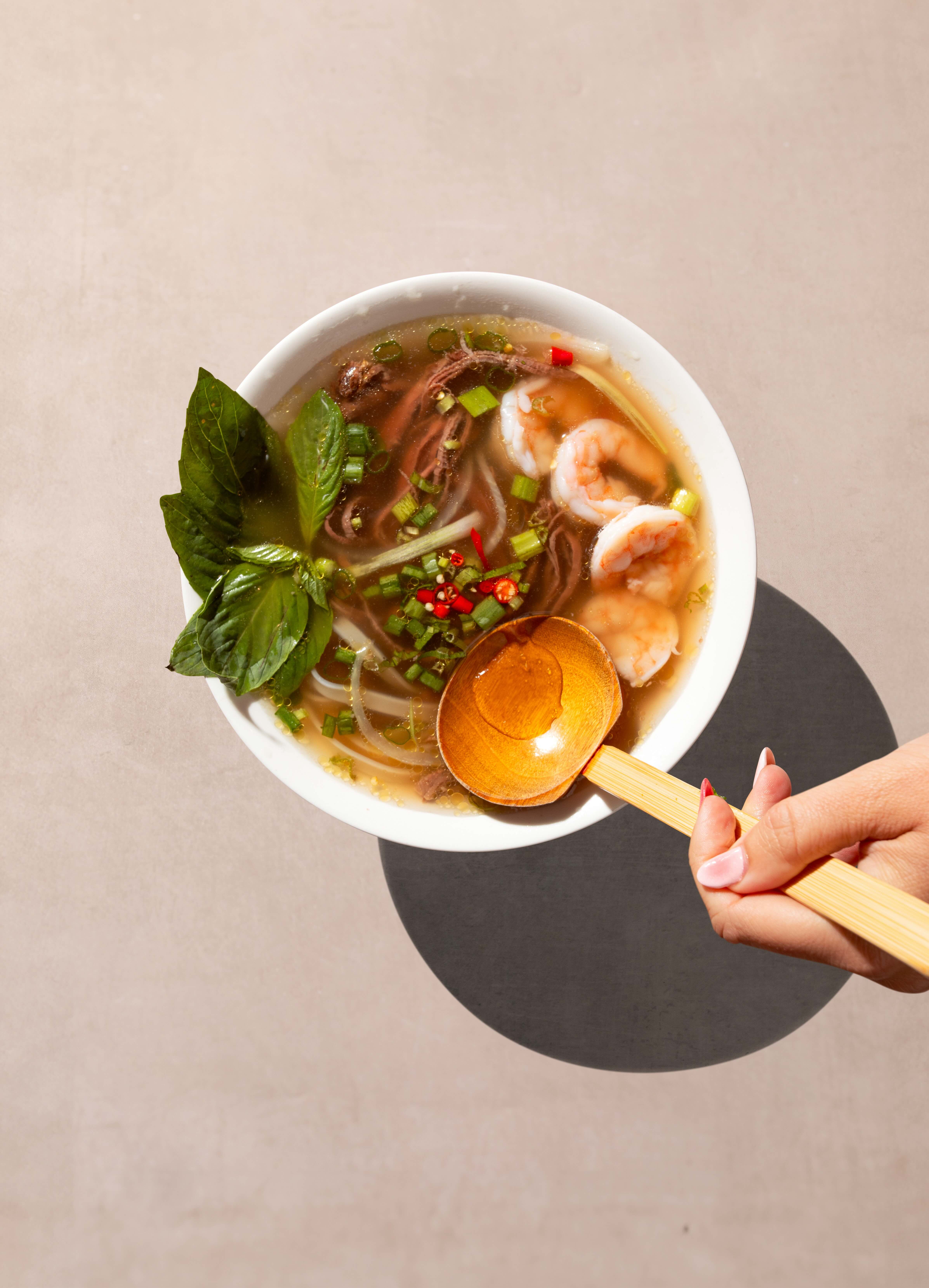 Hand holding wooden ladle over bowl of Vietnamese pho with prawns