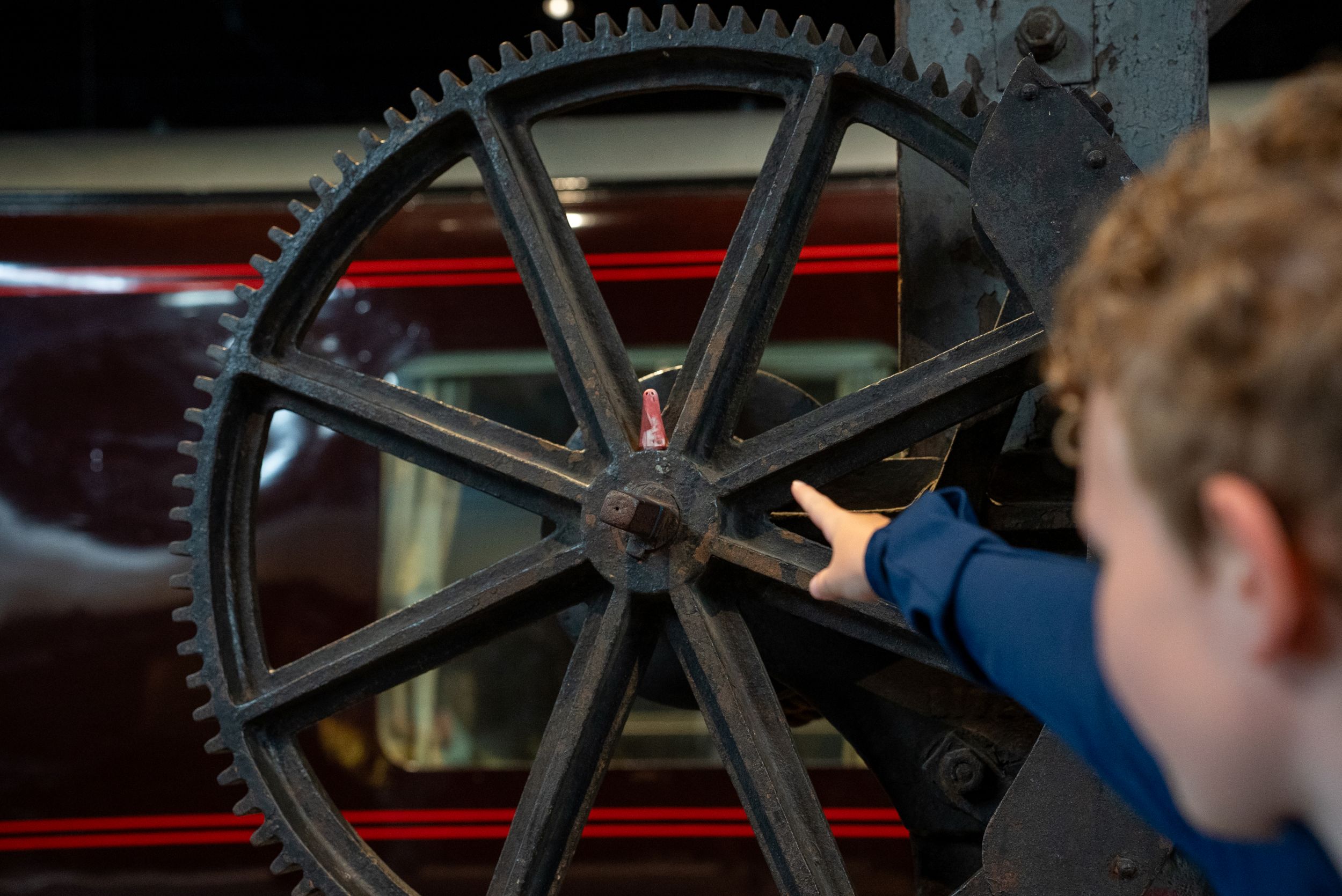 Child pointing at large industrial gear wheel with red train in background
