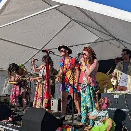 Colorful band performing on outdoor stage under white tent