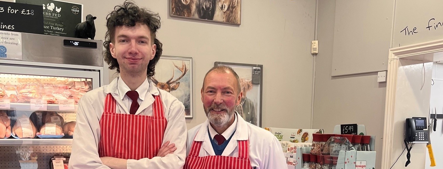 Two butchers in red aprons smiling inside a traditional shop