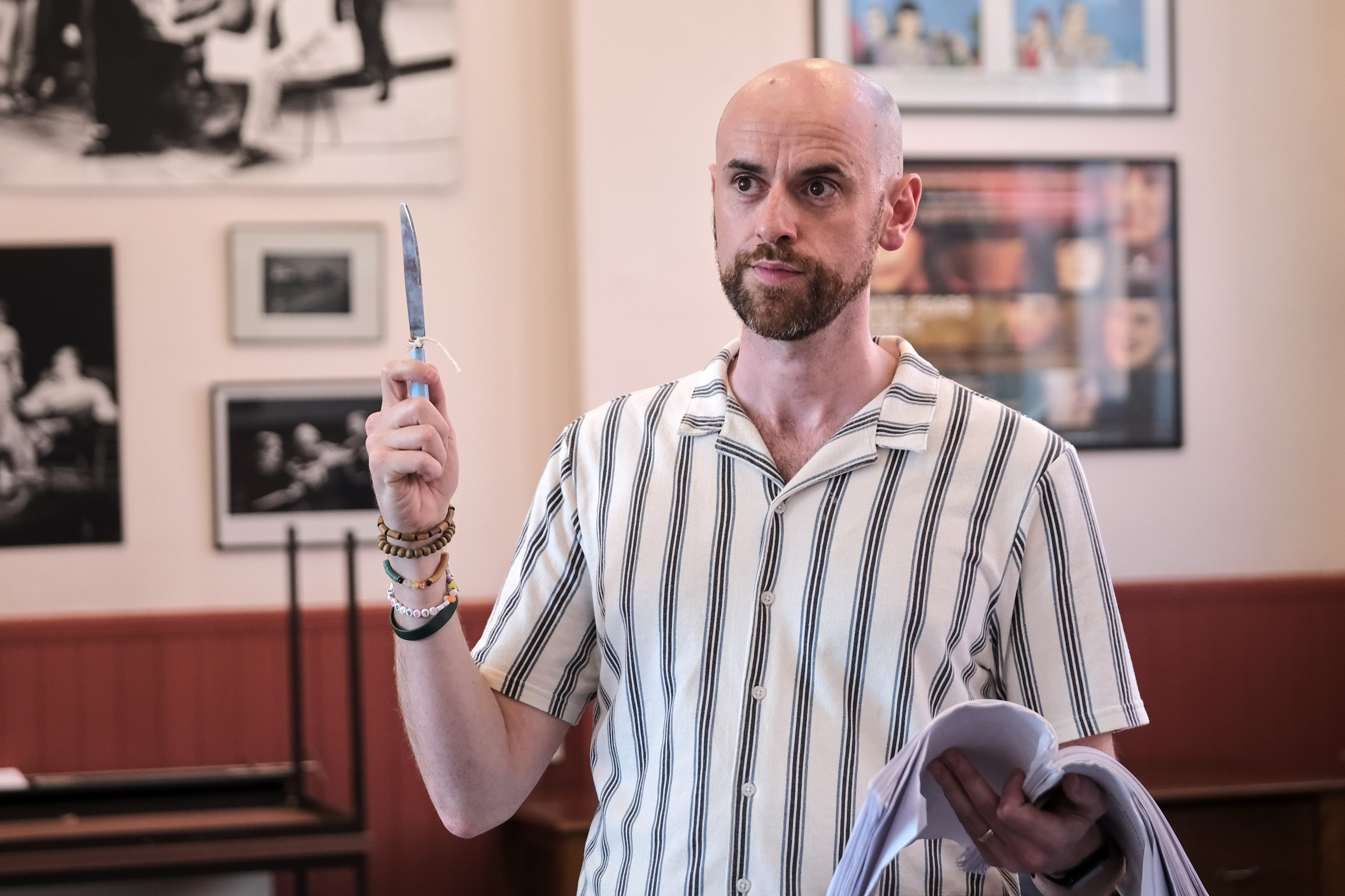 Bald man in striped shirt holding a knife in a room with photos