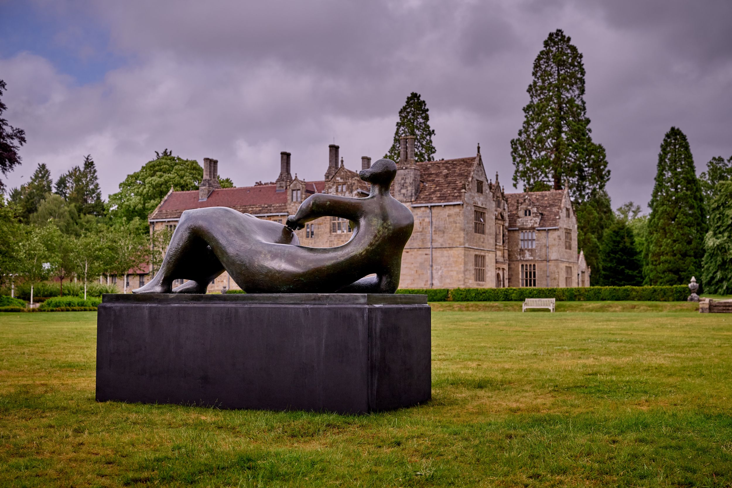 Sculpture of reclining figure on lush lawn with historic manor