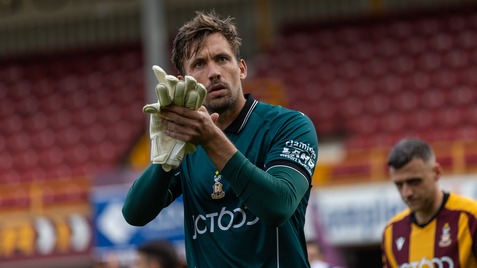 Football player applauding in green jersey with gloves on pitch