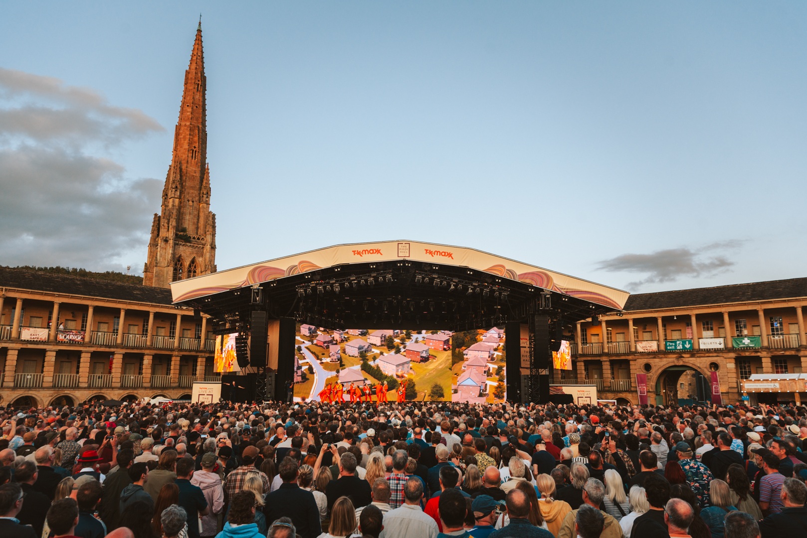 Large crowd at outdoor concert with tall spire and lit stage