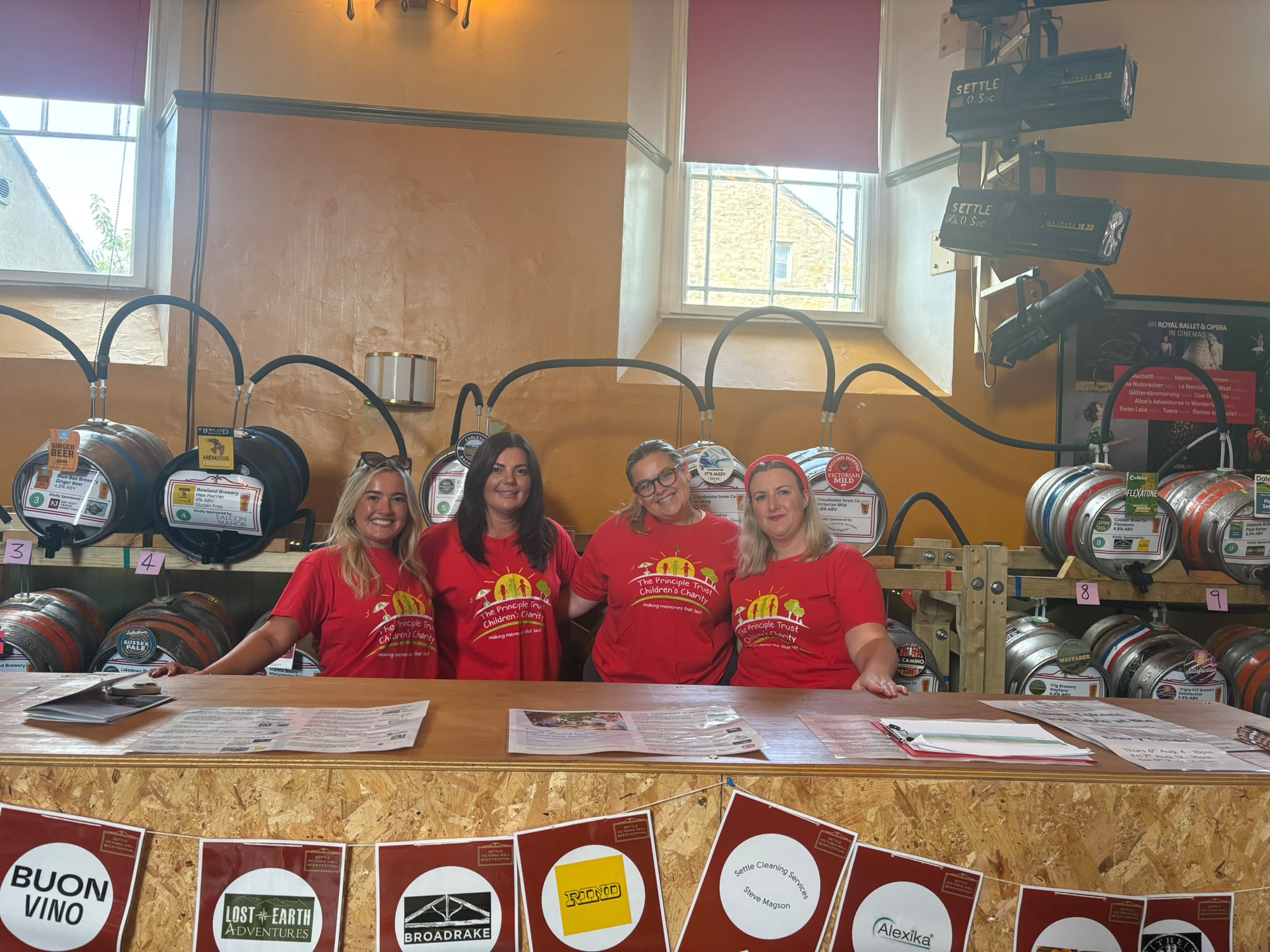 Four women in red shirts behind a bar with beer kegs