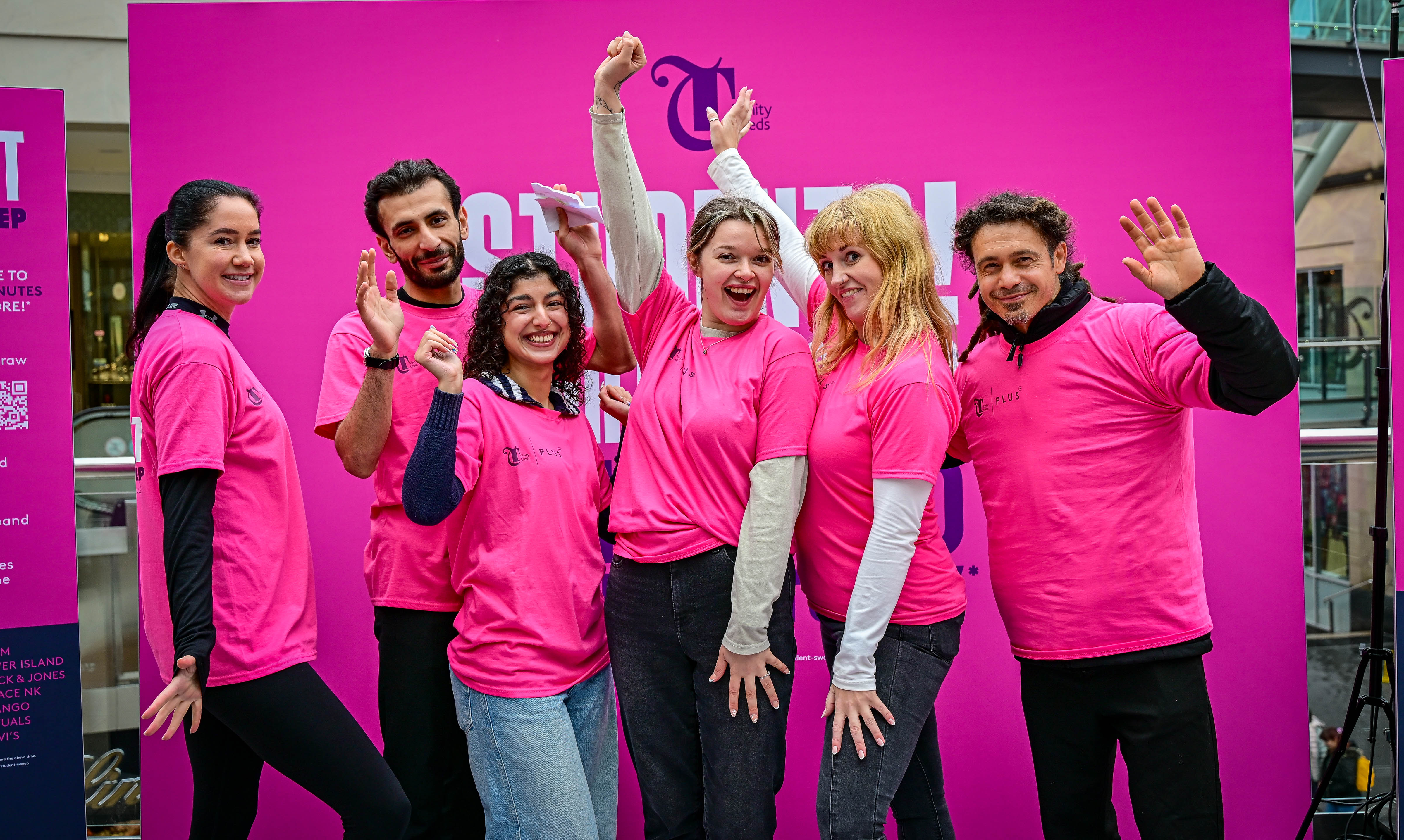 Group of six people in pink shirts smiling and waving indoors