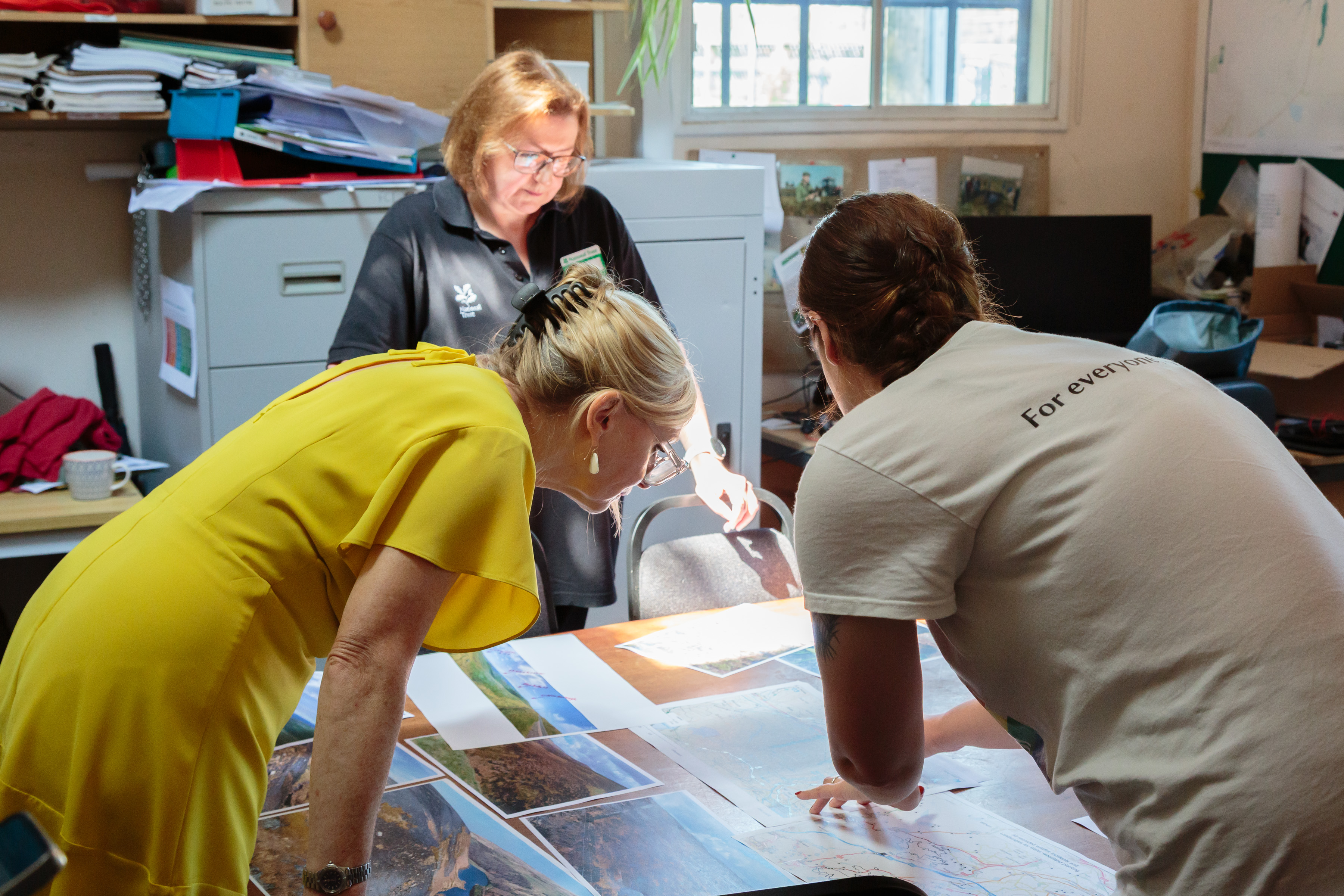 Three women examining maps on a table in a sunlit office
