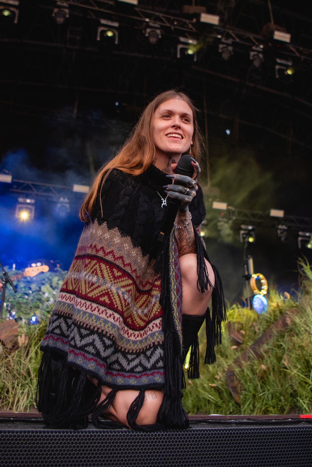Person with long hair kneeling on stage wearing patterned poncho