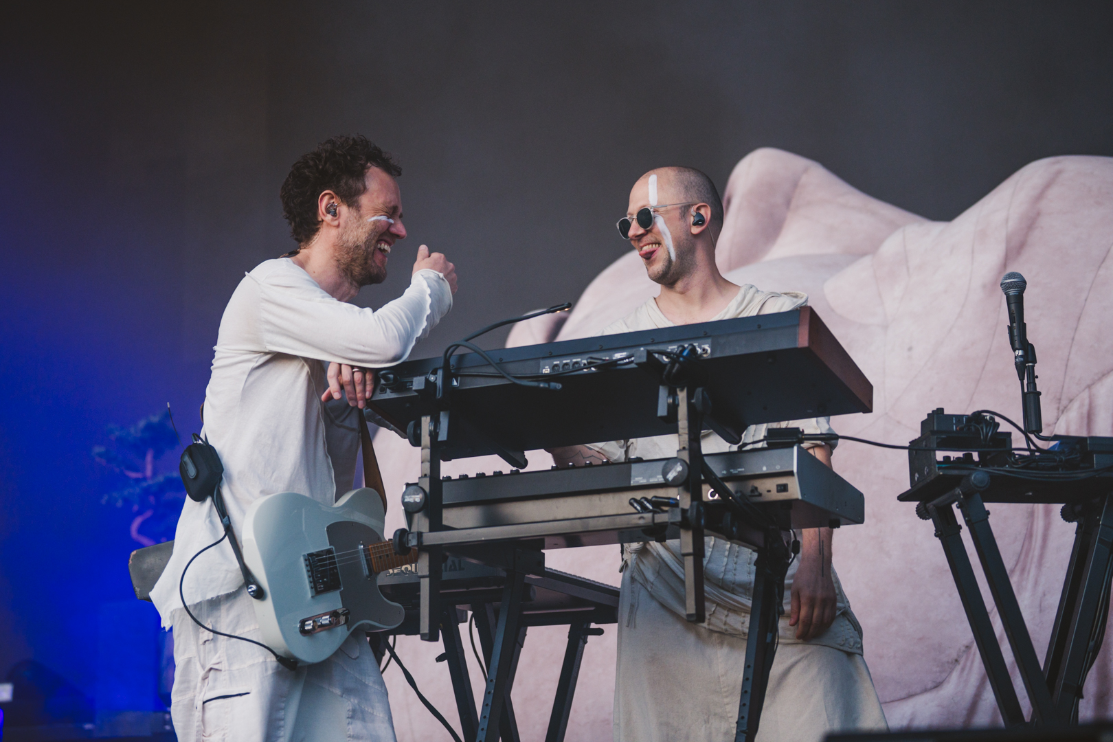 Two musicians in white outfits laughing on stage with keyboards