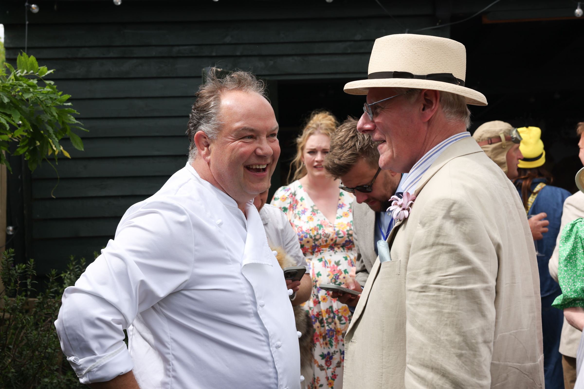 Chef laughing with a man in a beige suit and straw hat outdoors