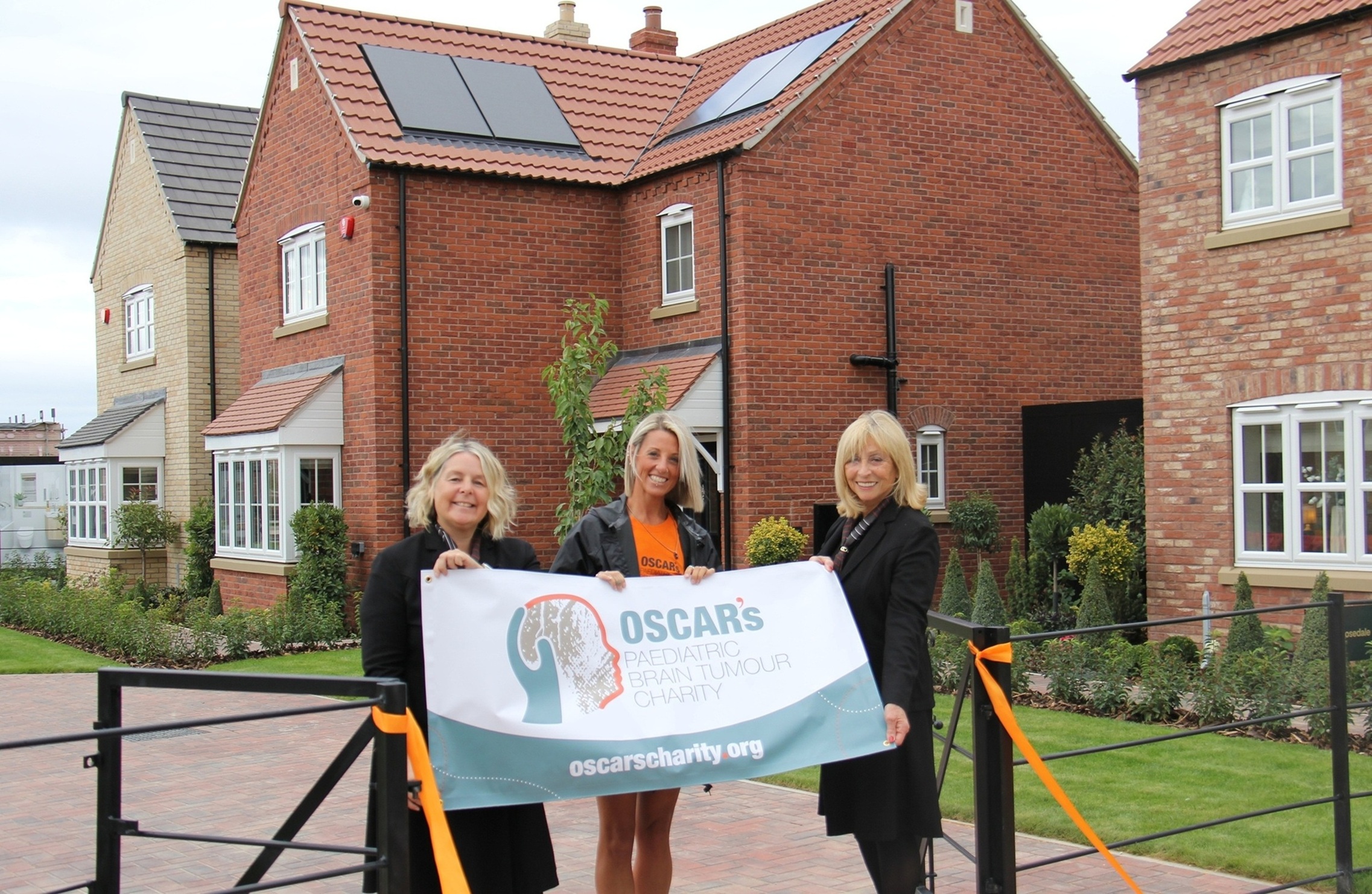 Three women holding a charity banner in front of new brick houses