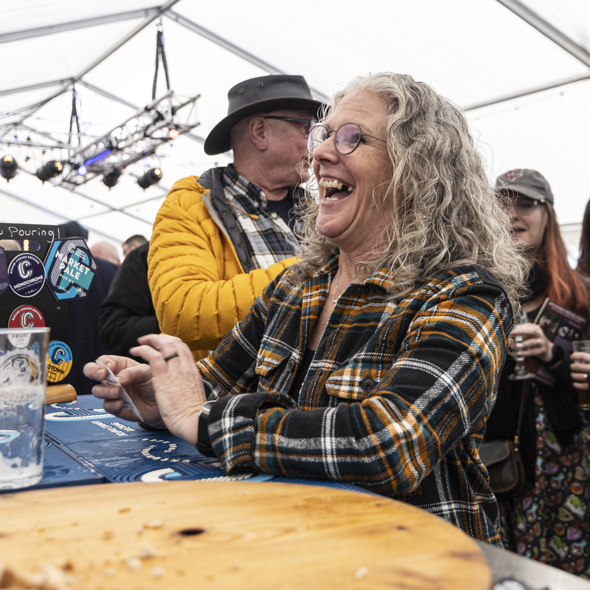 Smiling woman in a plaid shirt at a lively beer festival tent