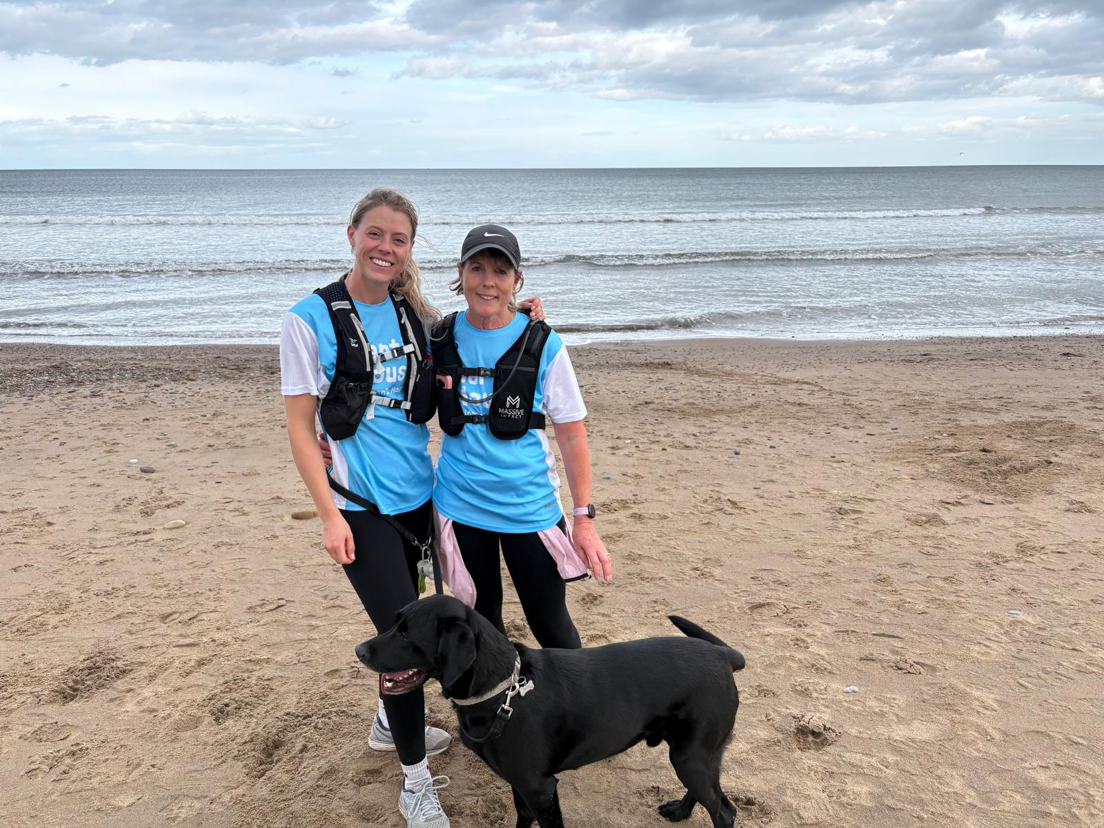 Two women in blue vests with a black dog on a sandy beach