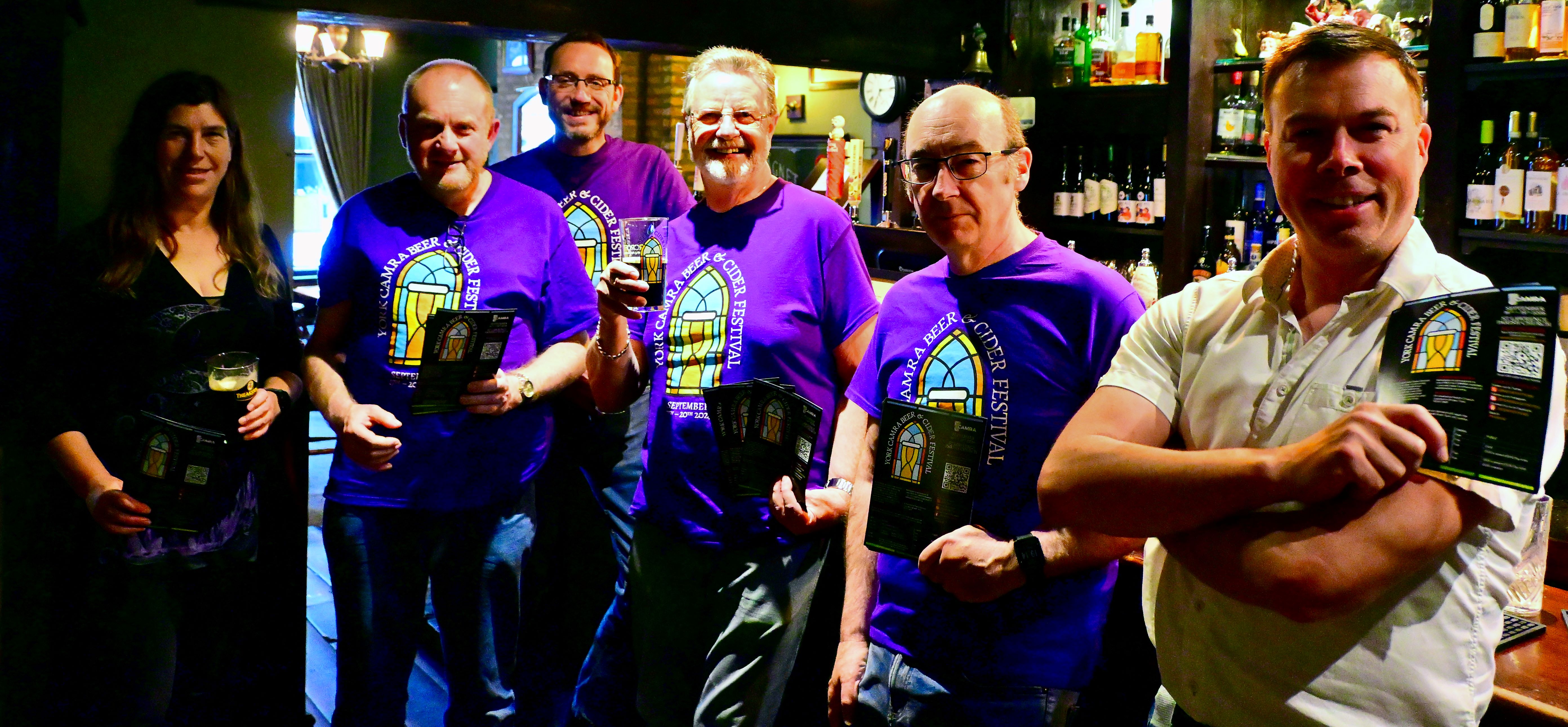 Group of people in a pub wearing purple festival shirts holding flyers