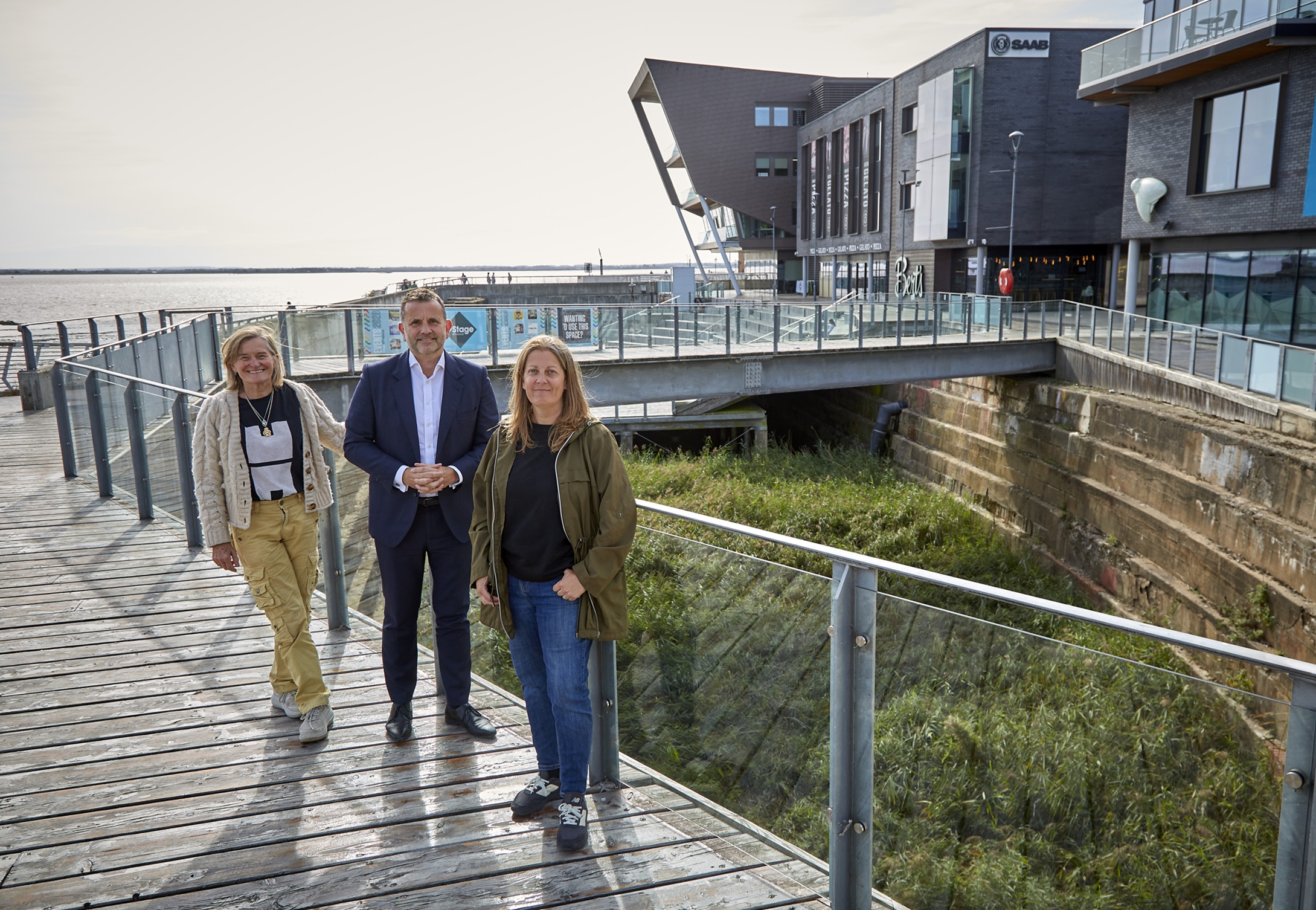 Three people on wooden boardwalk beside modern riverside buildings