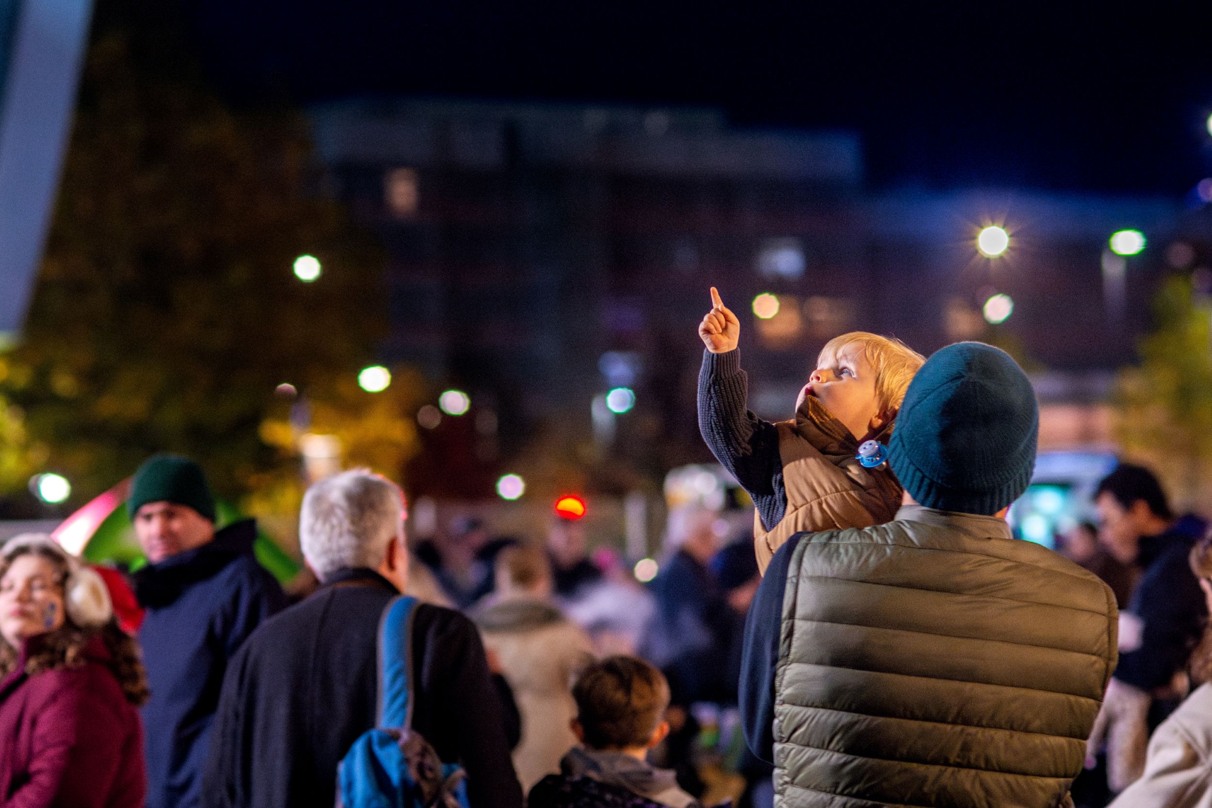 Child on adult's shoulders pointing at night sky during city event