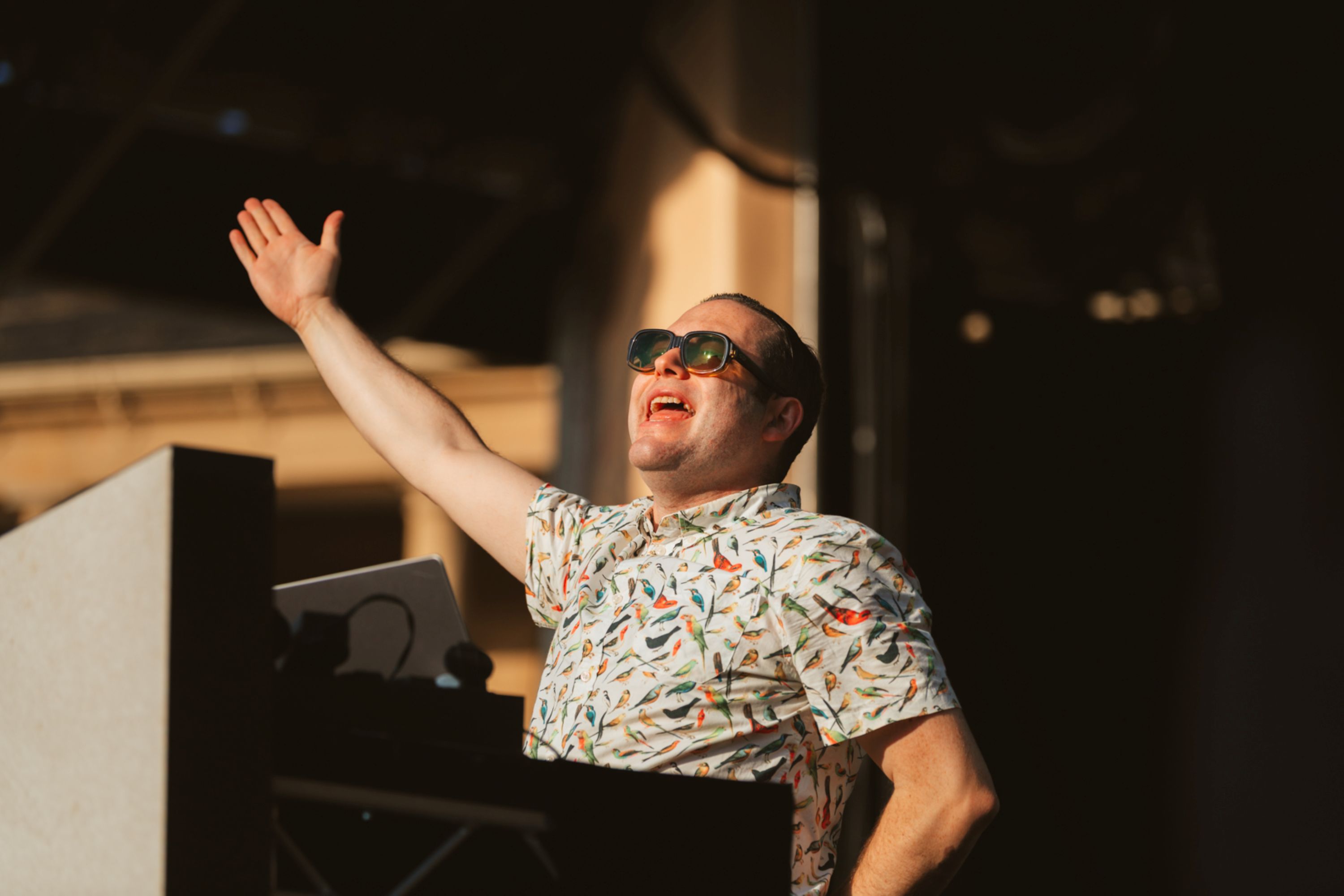 Man in floral shirt and sunglasses speaking passionately on stage