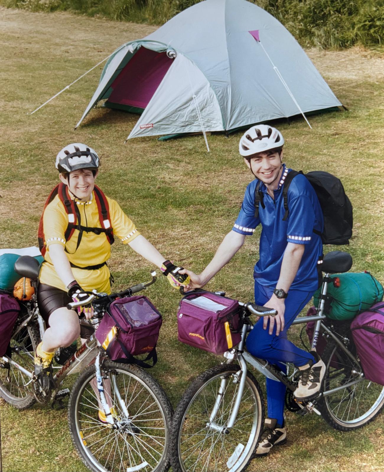 Two cyclists in colourful gear with loaded bikes near a tent