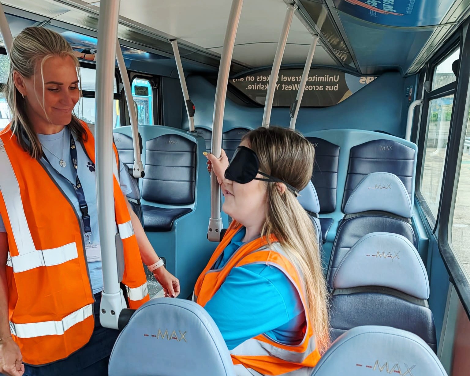 Inside a bus, a person wearing a high-visibility orange vest and a vision-impairment simulation blindfold is seated and holding a grab pole while taking part in an accessibility awareness exercise. Another person in a high-visibility vest stands nearby, providing guidance. The bus interior features blue seats, handrails, and large windows.