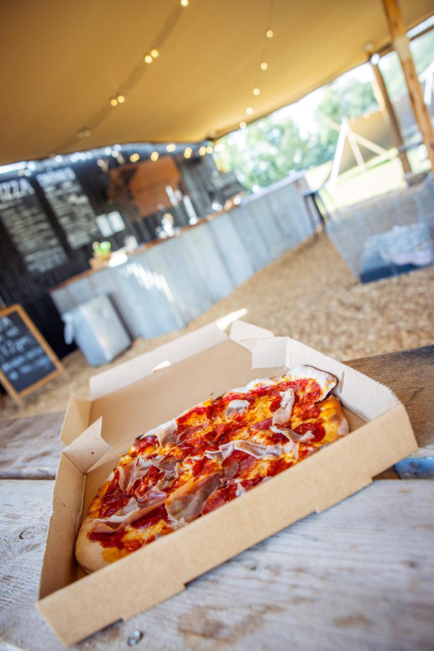 Wood-fired pizza in box at rustic outdoor eatery with menu board