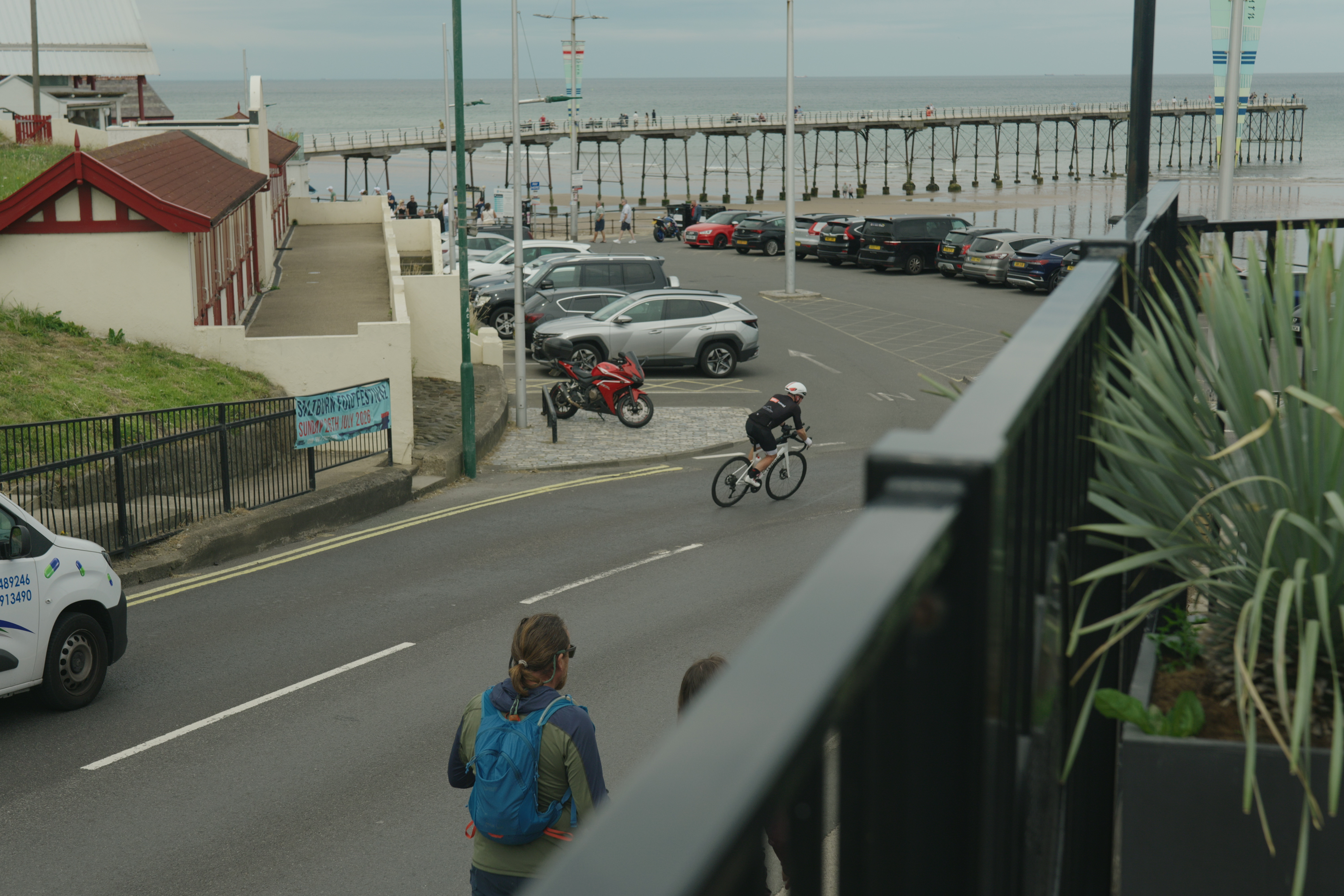 Cyclist on coastal road near pier with parked cars