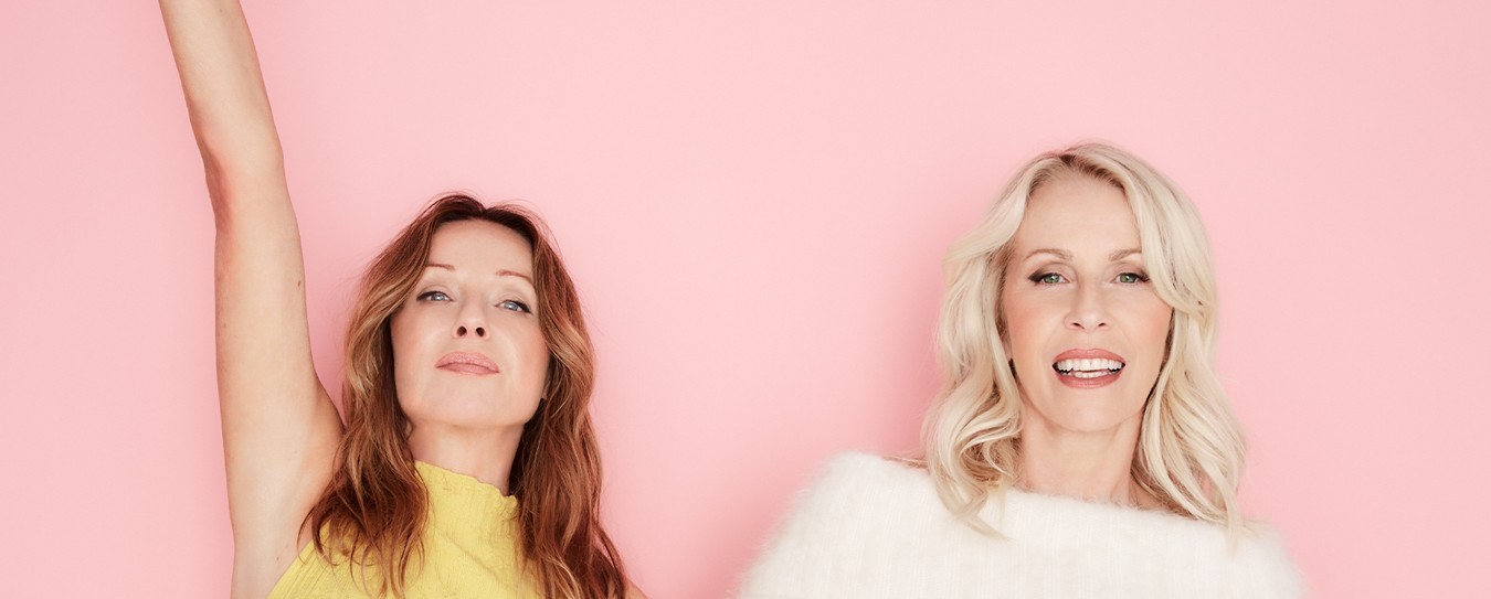 Two smiling women against a pastel pink background, one with arm raised
